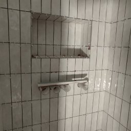 Multiservice - Świeżo ułożone, błyszczące, pionowe, białe płytki w łazience z wnęką na kosmetyki i mocowaniem na akcesoria, widoczne krzyżyki dystansowe.