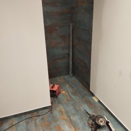 Multiservice - Łazienka w trakcie remontu: ułożone płytki imitujące rdzę na podłodze i ścianach, widoczny narożnik, narzędzia glazurnicze - szlifierka kątowa i pistolet do silikonu, oraz spawarka inwertorowa.