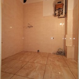 Multiservice - Jasna łazienka w trakcie prac wykończeniowych: ułożone płytki podłogowe i ścienne w odcieniach beżu, zdemontowany piecyk gazowy, widoczne ślady po instalacji i brakujące elementy wyposażenia.