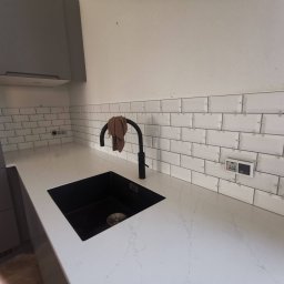 Multiservice - Białe, prostokątne płytki ułożone na ścianie nad blatem z czarnym zlewem i baterią. Widoczne krzyżyki dystansowe między płytkami, sugerujące etap prac. Szare szafki kuchenne w tle.