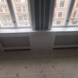 Multiservice - Biała zabudowa grzejnika pod oknem na poddaszu, widok z dołu na drewnianą podłogę i budynki za oknem.