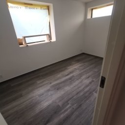 Multiservice - Wnętrze pokoju z ułożoną podłogą z paneli imitujących drewno, okno zabezpieczone folią i drewnianą konstrukcją, widok zza otwartych drzwi.