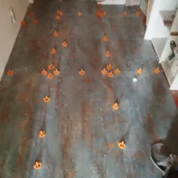 Podłoga z szarych płytek imitujących rdzę, z pomarańczowymi klipsami poziomującymi i cienkimi sznurkami do fugowania, widoczne fragmenty listew przypodłogowych i drzwi.