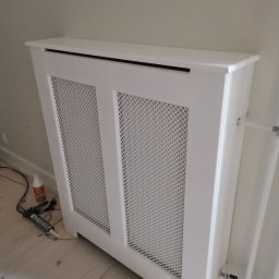 Multiservice - Biała, ażurowa obudowa grzejnika zamontowana przy ścianie, widoczne narzędzia montażowe i kabel zasilający na podłodze.