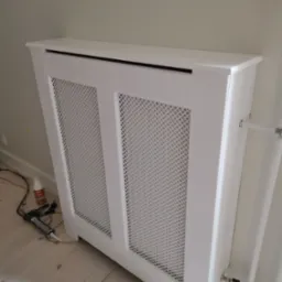 Biała, ażurowa obudowa grzejnika zamontowana przy ścianie, widoczne narzędzia montażowe i kabel zasilający na podłodze.