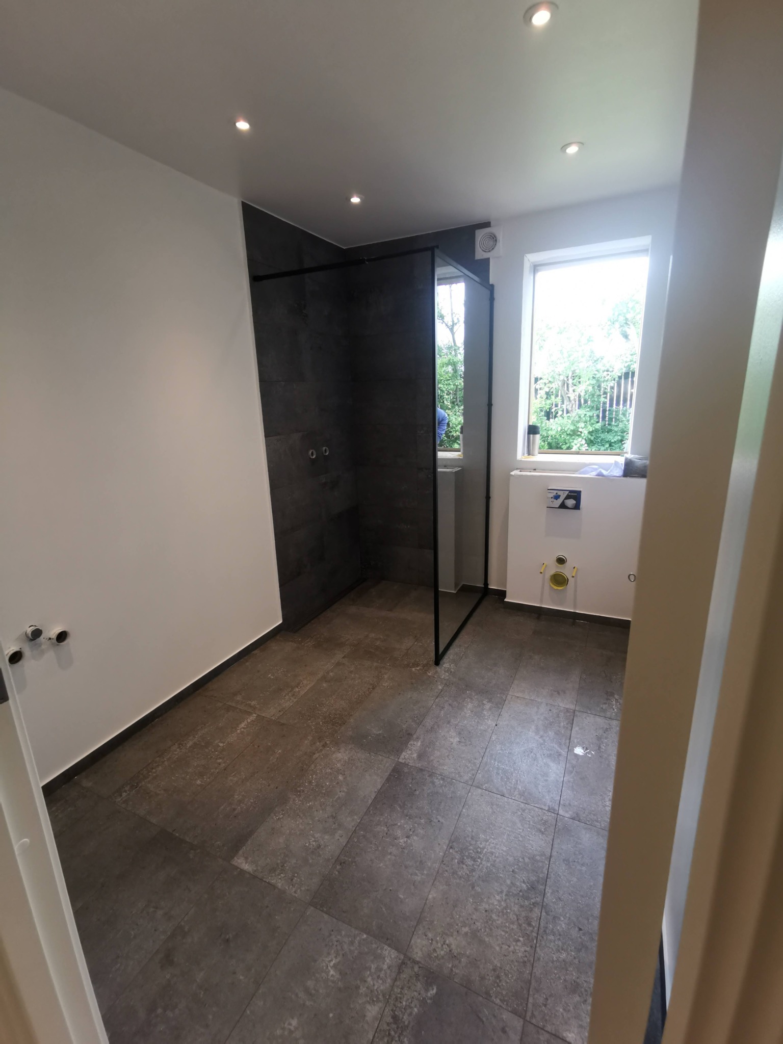 Łazienka w trakcie remontu: widoczna kabina prysznicowa typu walk-in z czarnymi ramami, szare płytki podłogowe i ścienne, przygotowane przyłącza wodno-kanalizacyjne, oraz zabudowa podtynkowego WC...