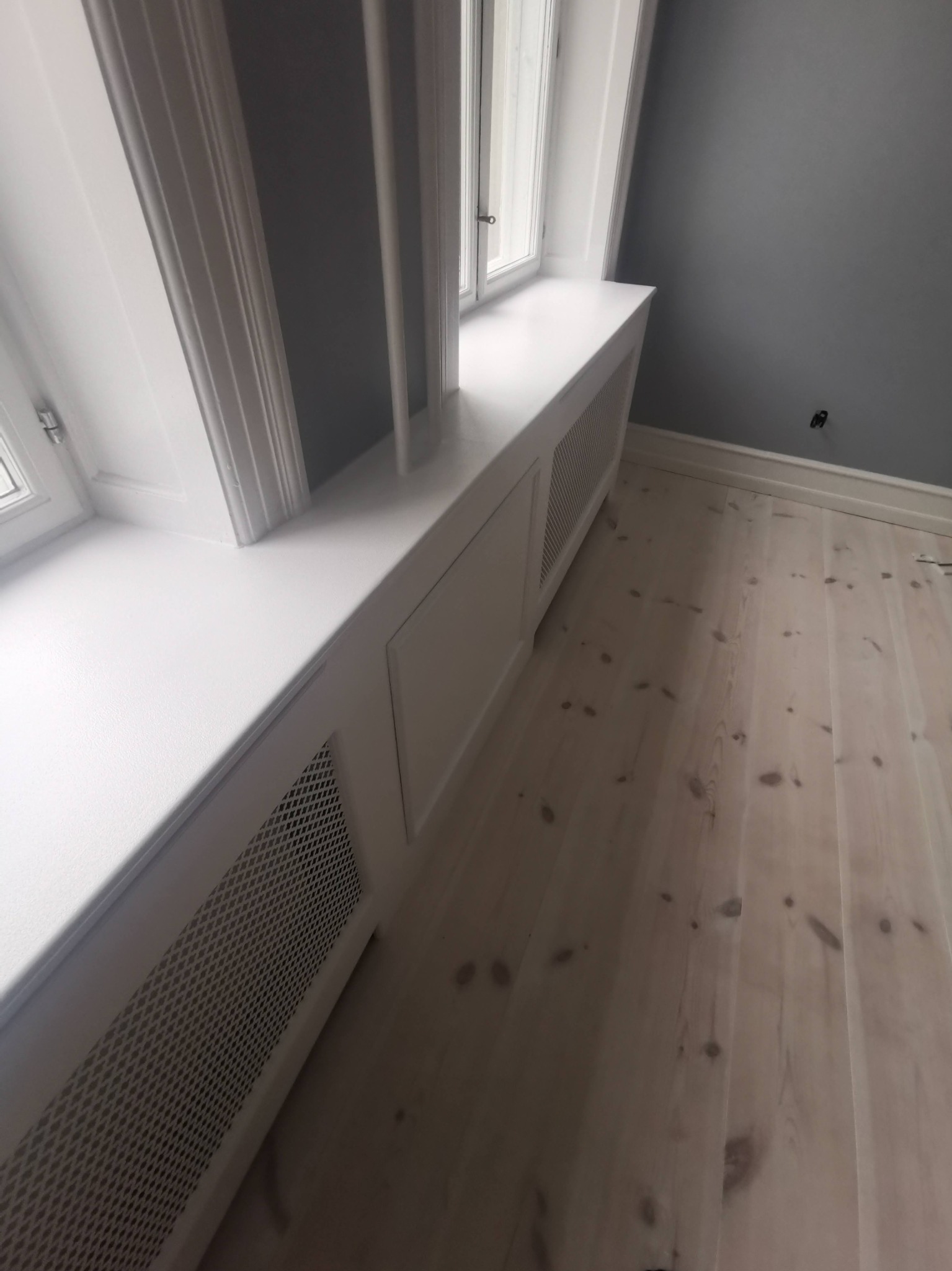 Biały, zabudowany grzejnik pod oknem w Tarnobrzegu, z drewnianą podłogą i szarą ścianą w tle.