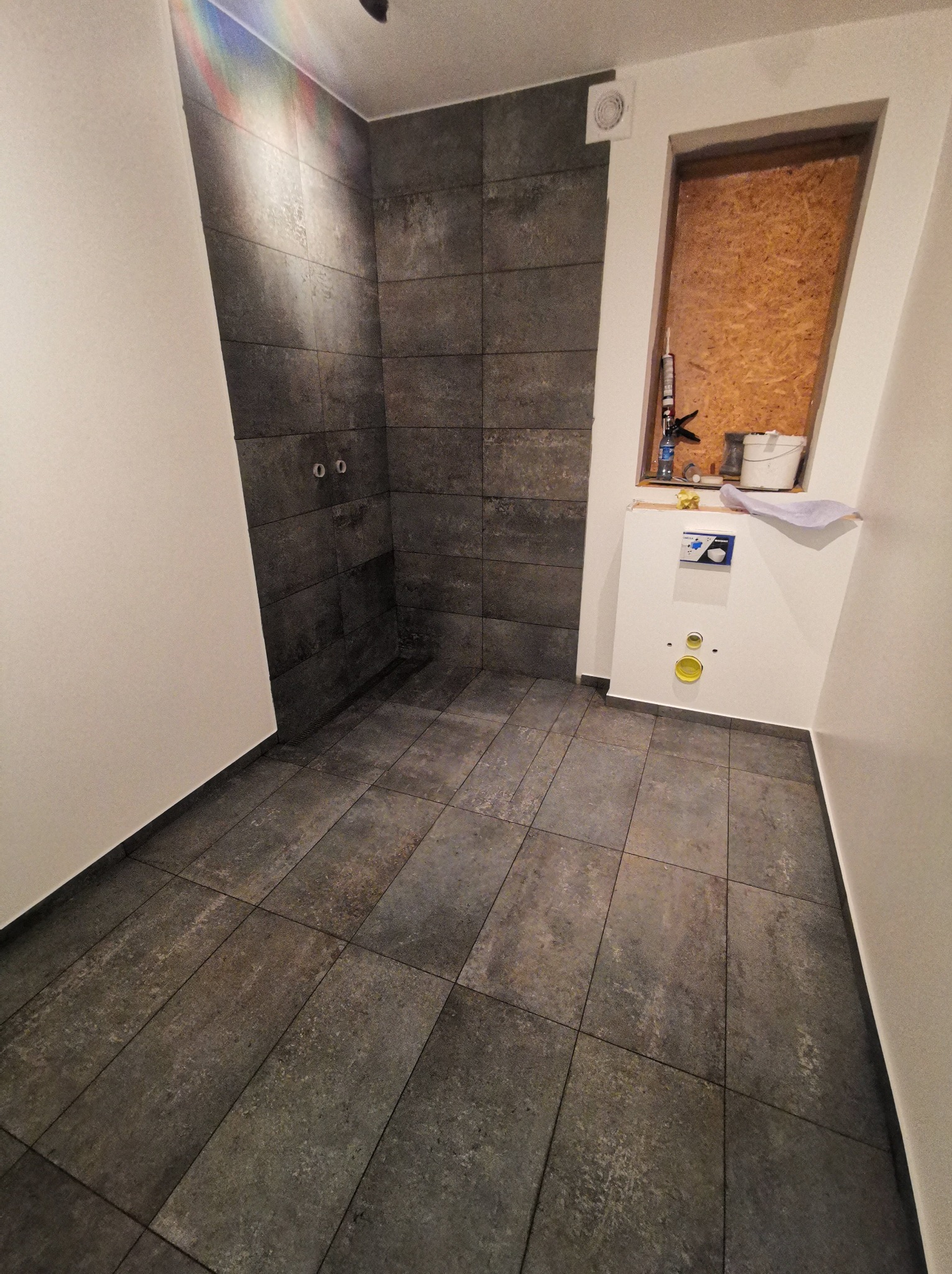 Wnętrze łazienki w trakcie prac wykończeniowych, widoczna ściana wyłożona ciemnymi płytkami imitującymi kamień, przygotowane przyłącza, zamontowany stelaż podtynkowy WC, oraz narzędzia i materiały...