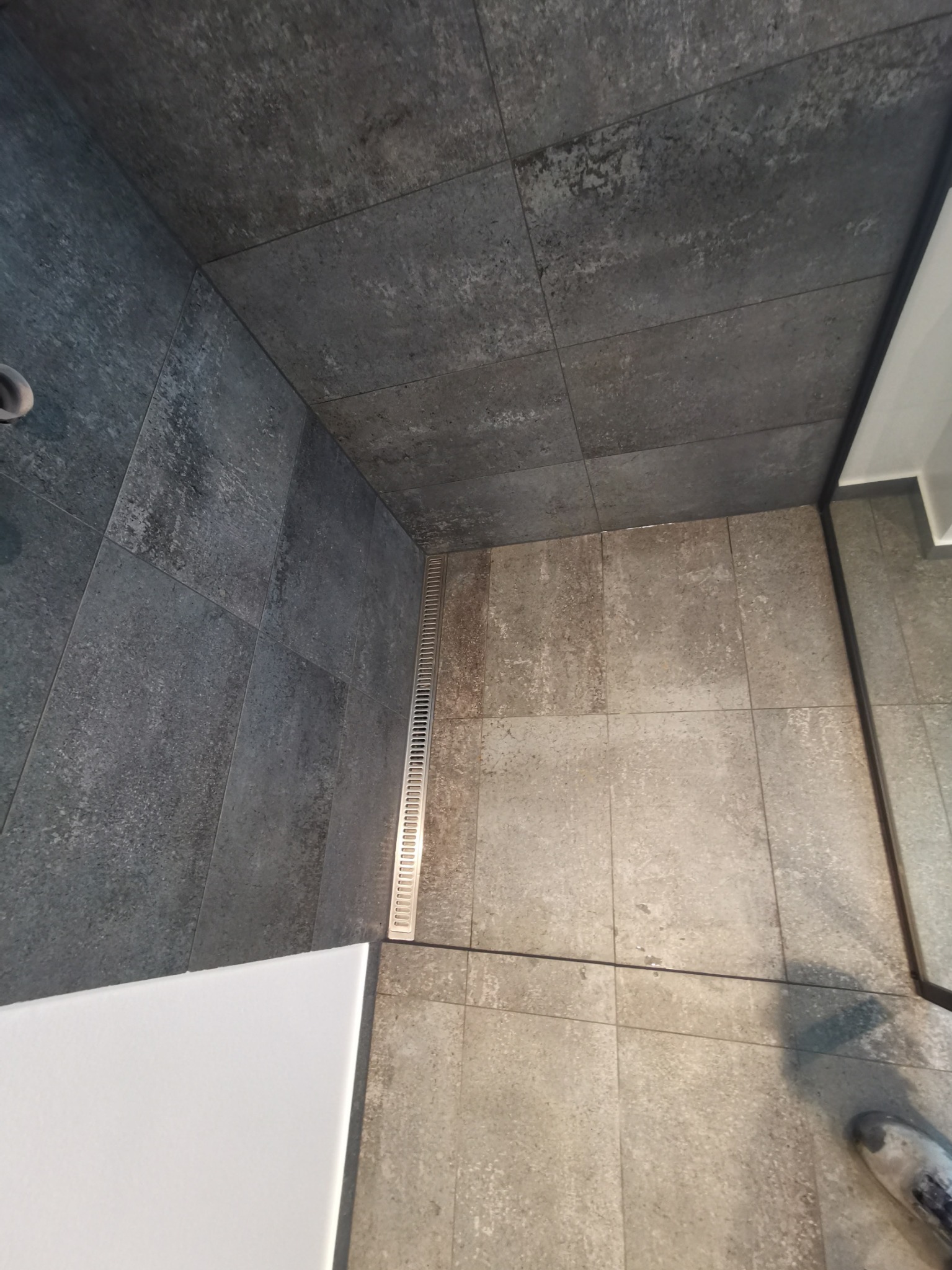 Nowoczesna kabina prysznicowa z szarymi płytkami imitującymi beton na ścianach i podłodze, z odpływem liniowym i fragmentem białego blatu umywalki.