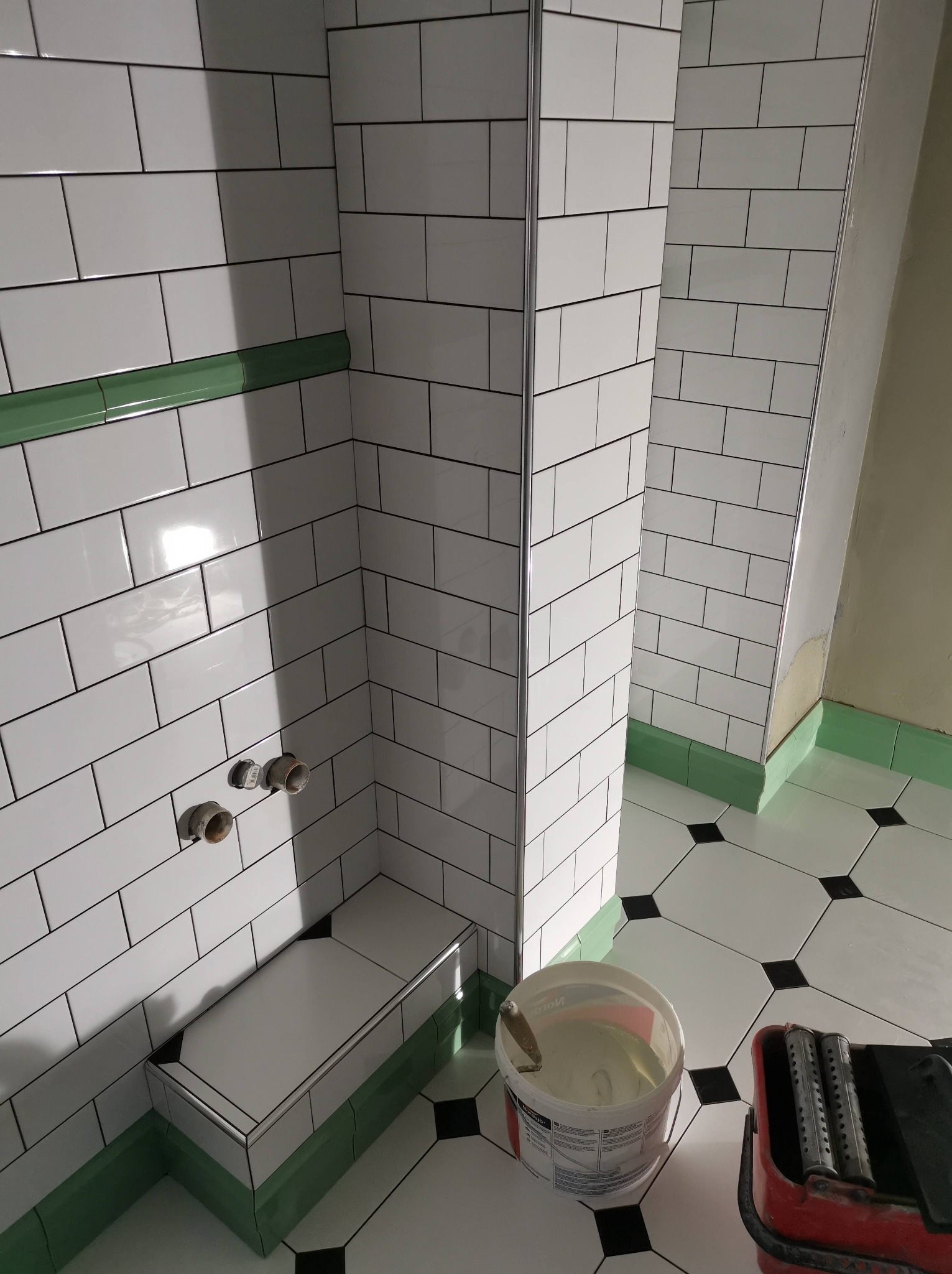 Białe, prostokątne płytki ułożone na ścianach i podłodze w łazience, z zielonymi akcentami na listwach przypodłogowych i obramowaniach. Widoczne wystające rury instalacji wodnej na ścianie i wiadro...