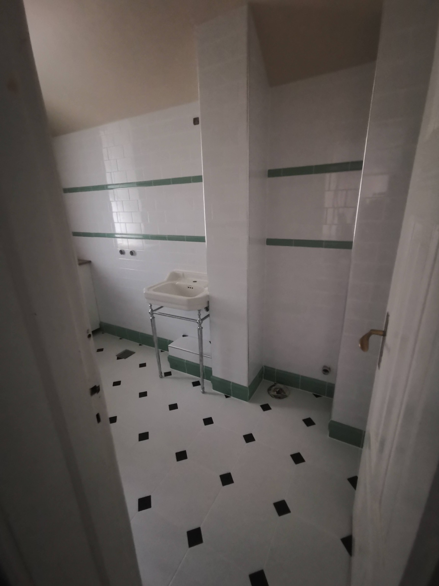 Biała łazienka z umywalką na chromowanych nogach, biało-czarną podłogą w kratkę i zielonymi akcentami na listwach przypodłogowych i pasie płytek na ścianie.