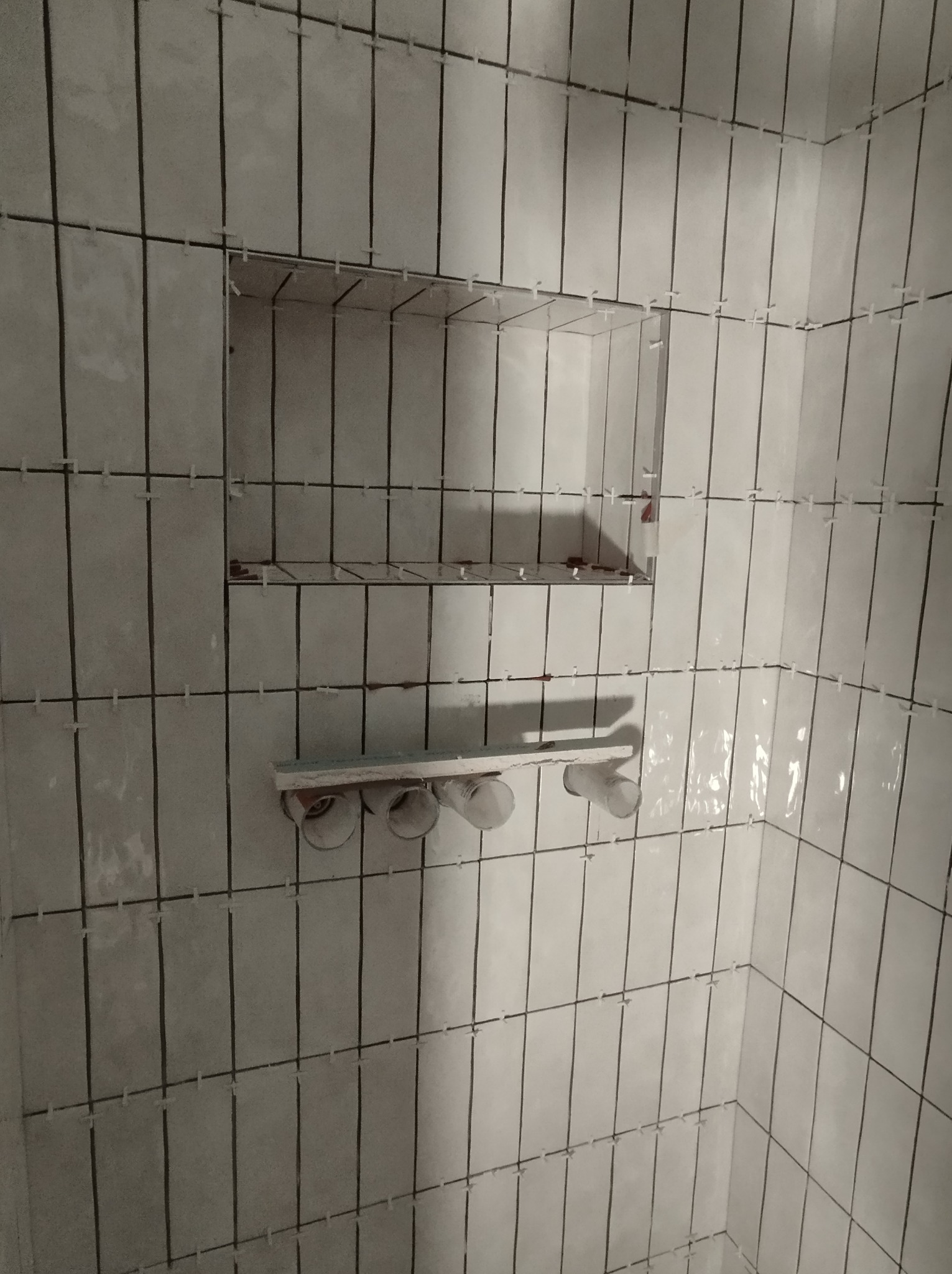 Świeżo ułożone, błyszczące, pionowe, białe płytki w łazience z wnęką na kosmetyki i mocowaniem na akcesoria, widoczne krzyżyki dystansowe.