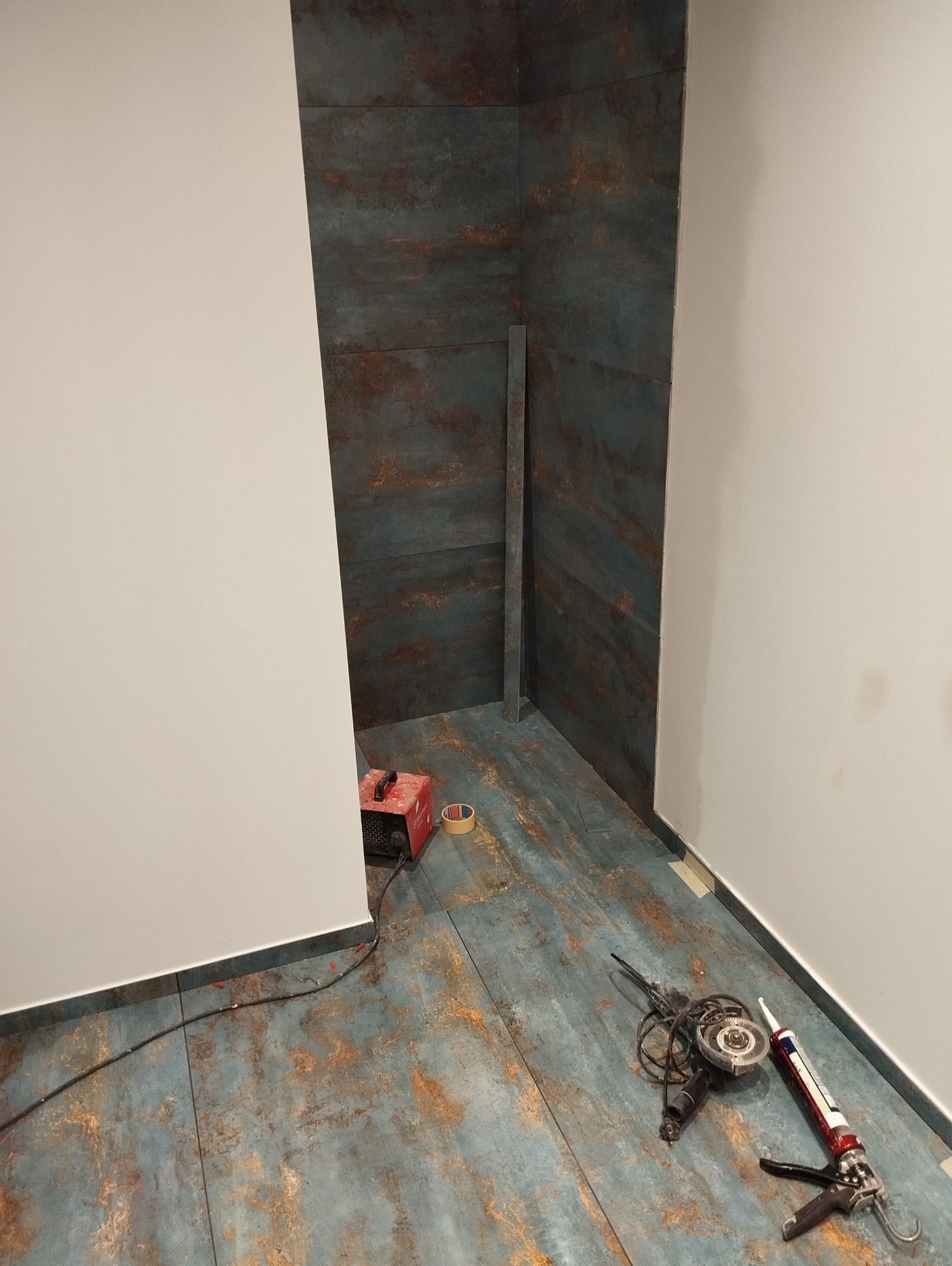 Łazienka w trakcie remontu: ułożone płytki imitujące rdzę na podłodze i ścianach, widoczny narożnik, narzędzia glazurnicze - szlifierka kątowa i pistolet do silikonu, oraz spawarka inwertorowa.
