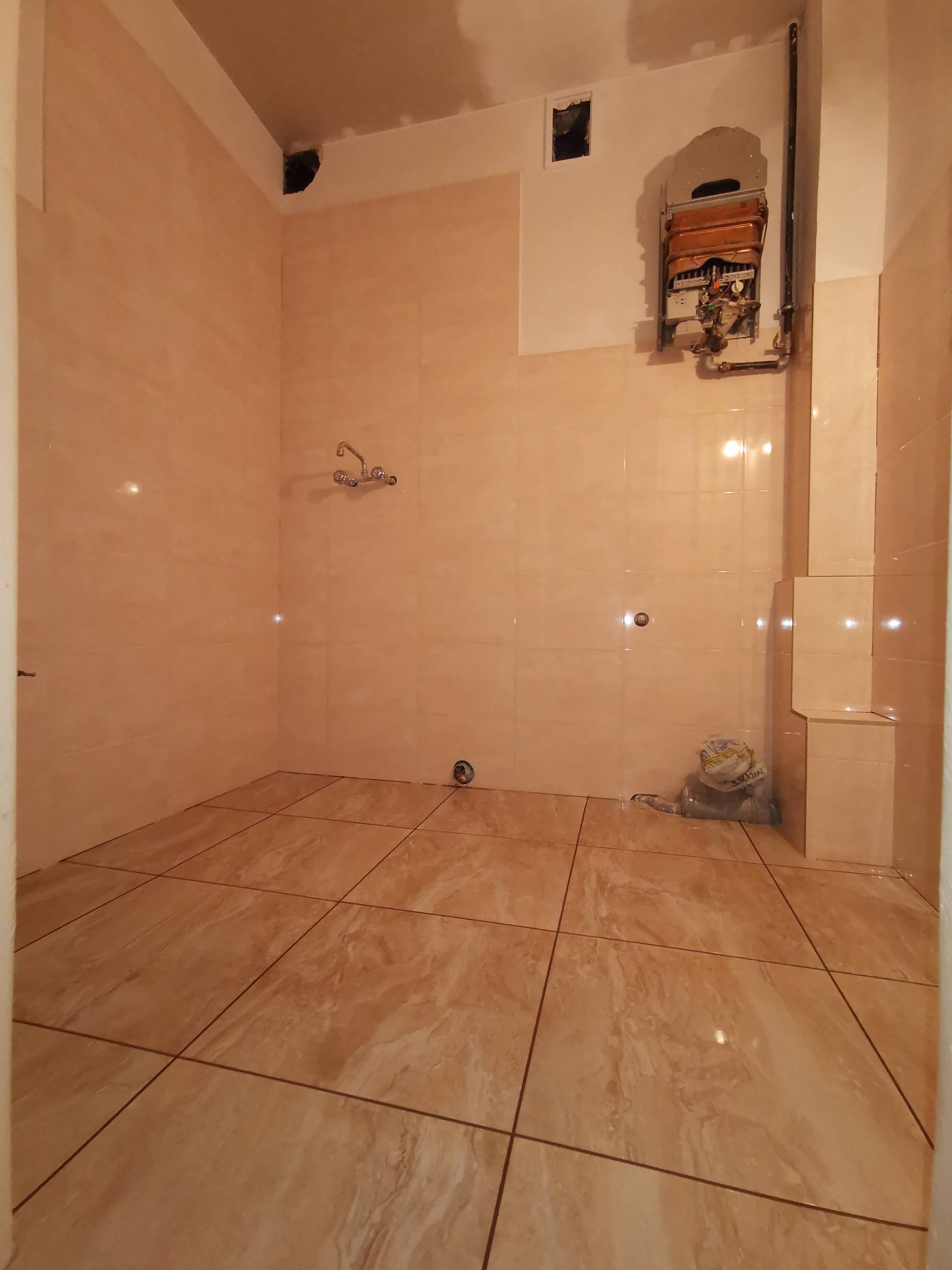 Jasna łazienka w trakcie prac wykończeniowych: ułożone płytki podłogowe i ścienne w odcieniach beżu, zdemontowany piecyk gazowy, widoczne ślady po instalacji i brakujące elementy wyposażenia.