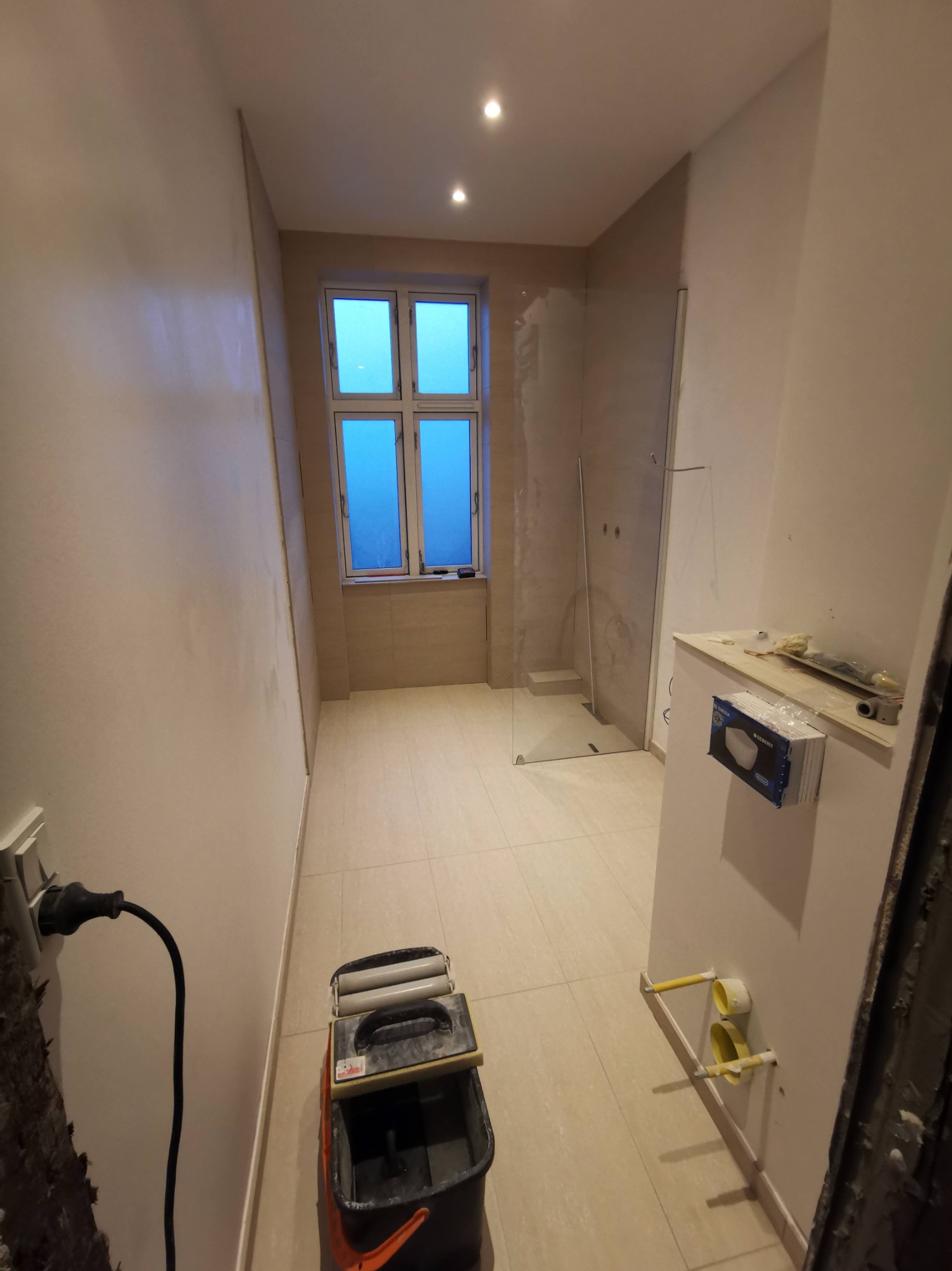 Łazienka w trakcie prac wykończeniowych: widoczna kabina prysznicowa ze szkła, okno, płytki na podłodze i ścianach, narzędzia glazurnicze oraz instalacja sanitarna w ścianie.