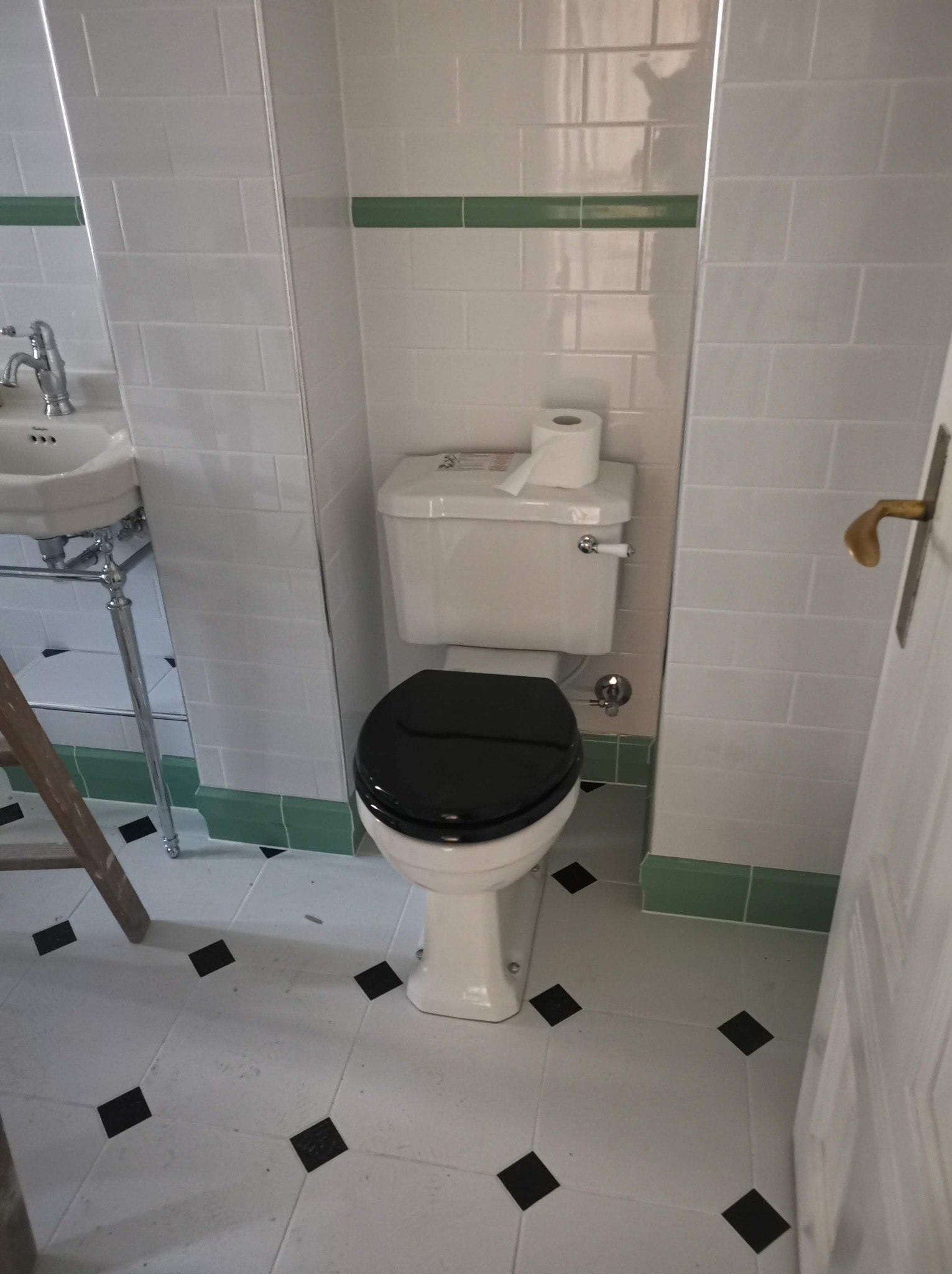 Toaleta z czarną deską sedesową, białą ceramiką i spłuczką, białe kafelki z zielonym paskiem dekoracyjnym, podłoga w biało-czarną szachownicę, widoczny fragment umywalki na chromowanych nogach...