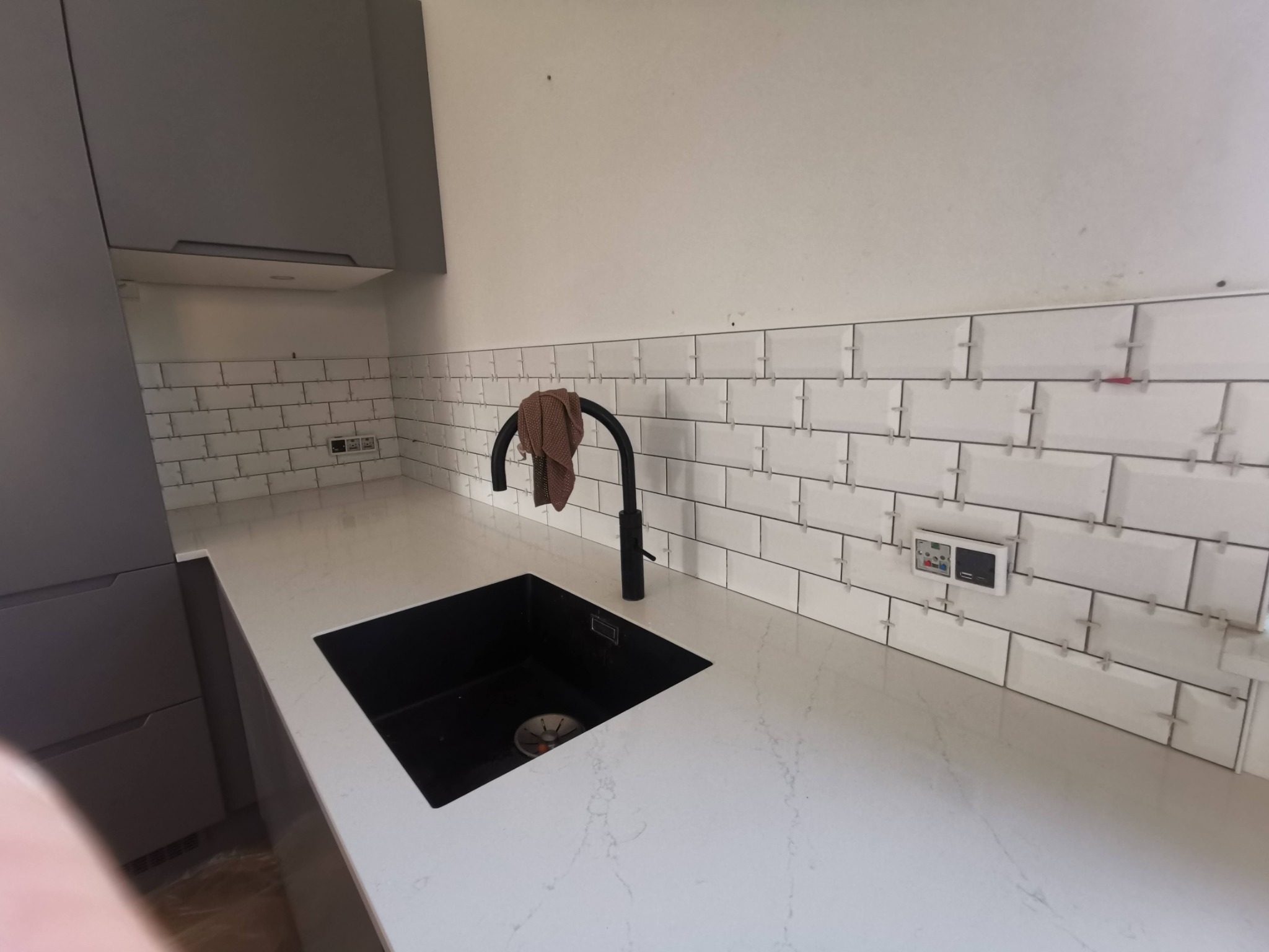 Białe, prostokątne płytki ułożone na ścianie nad blatem z czarnym zlewem i baterią. Widoczne krzyżyki dystansowe między płytkami, sugerujące etap prac. Szare szafki kuchenne w tle.