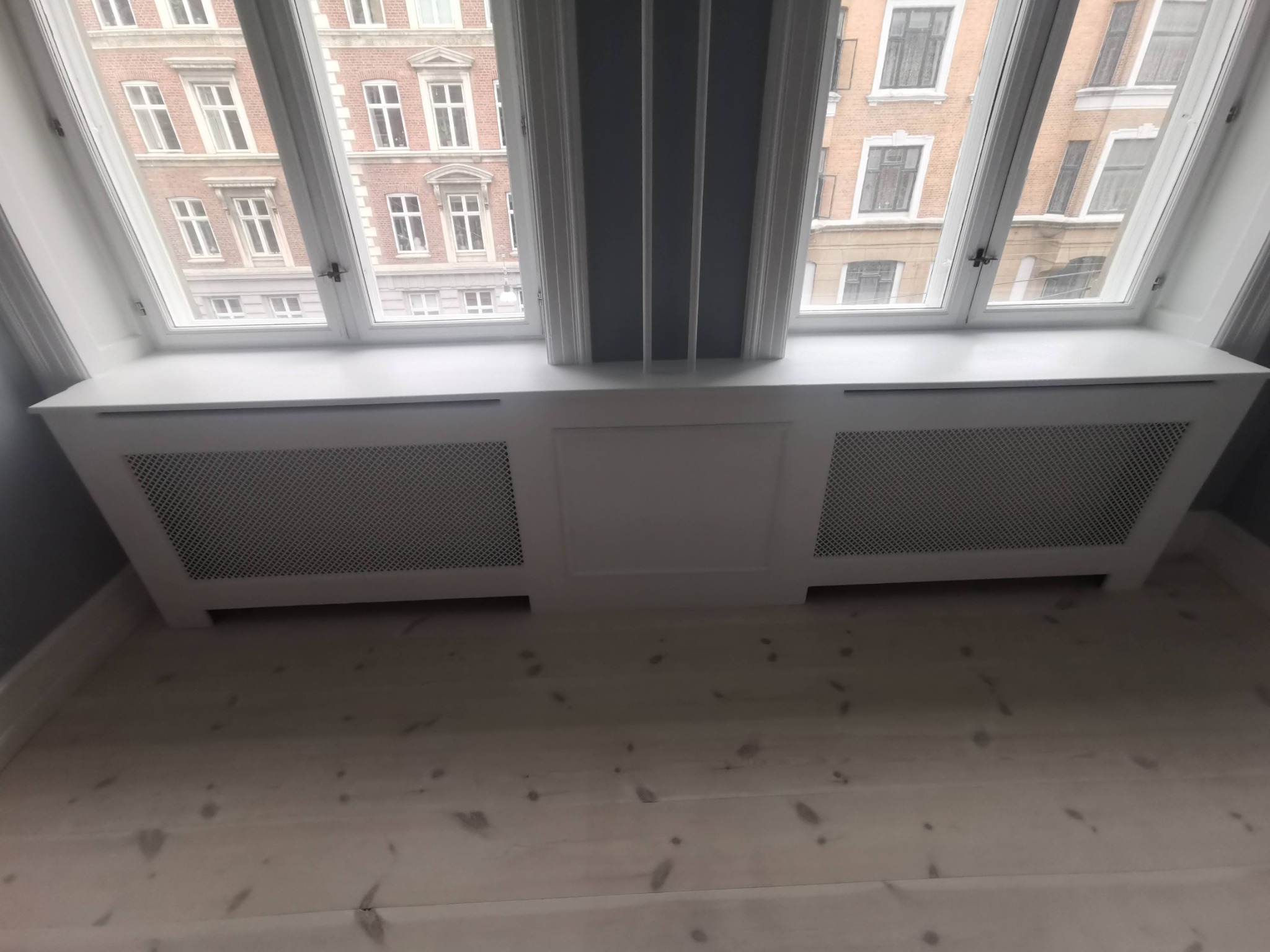 Biała zabudowa grzejnika pod oknem na poddaszu, widok z dołu na drewnianą podłogę i budynki za oknem.