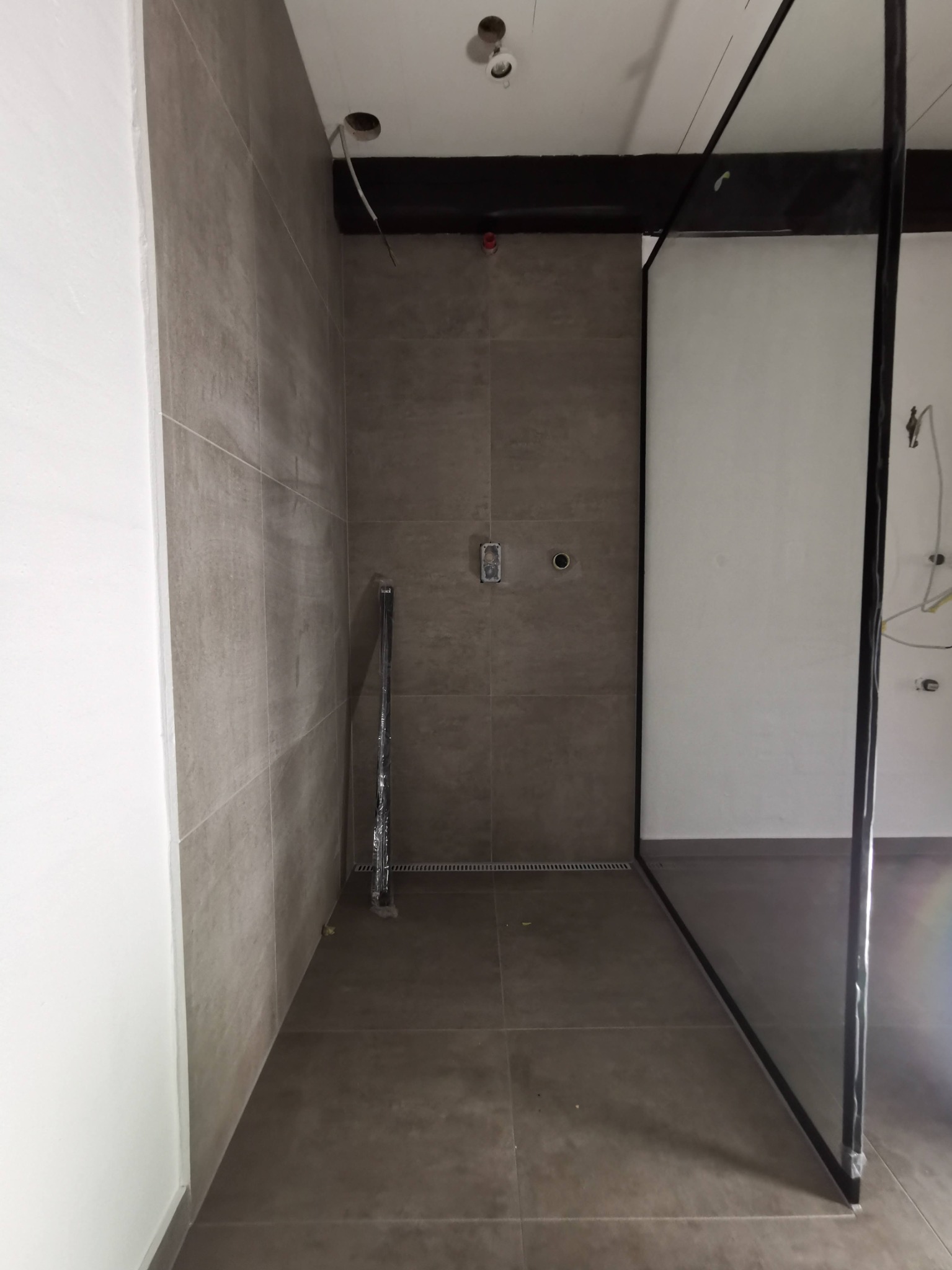 Przygotowana do montażu kabina prysznicowa typu walk-in z czarnymi profilami, szare płytki na ścianach i podłodze, widoczne instalacje elektryczne i wodne w surowym stanie.