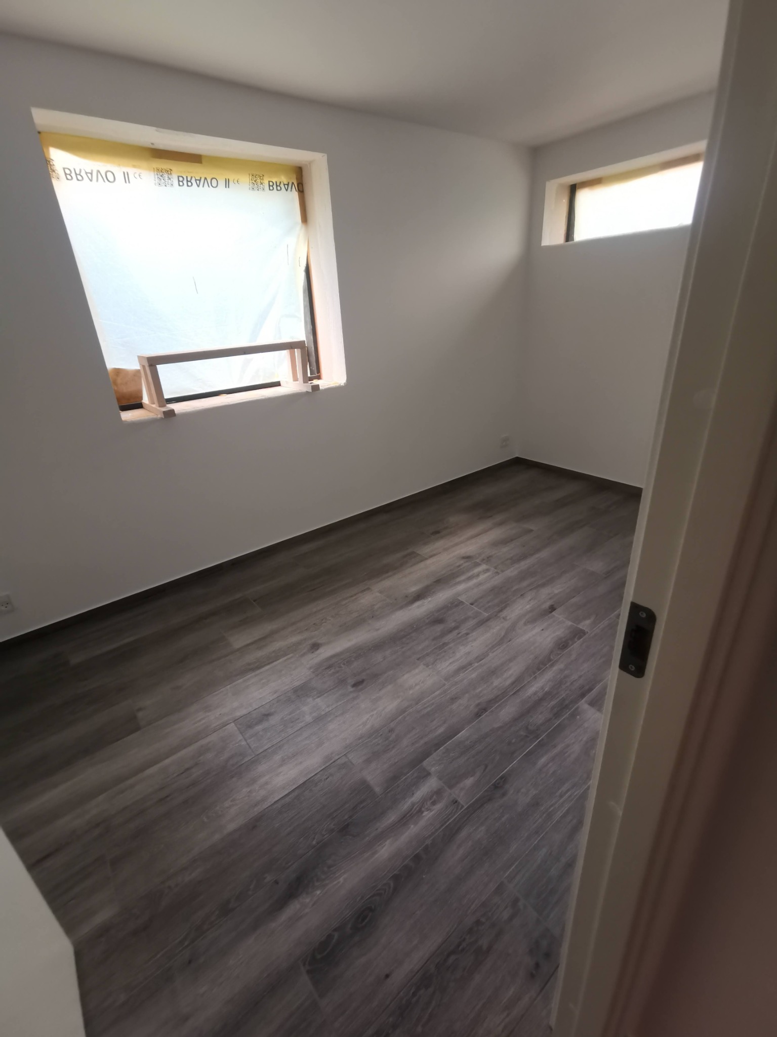 Wnętrze pokoju z ułożoną podłogą z paneli imitujących drewno, okno zabezpieczone folią i drewnianą konstrukcją, widok zza otwartych drzwi.