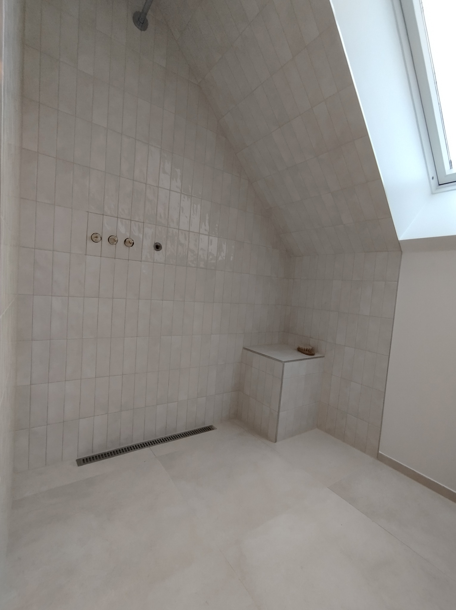 Wykończona kabina prysznicowa na poddaszu z beżowymi, podłużnymi płytkami, odpływem liniowym w podłodze i półką w rogu. Widoczne otwory na baterię i deszczownicę oraz okno dachowe.