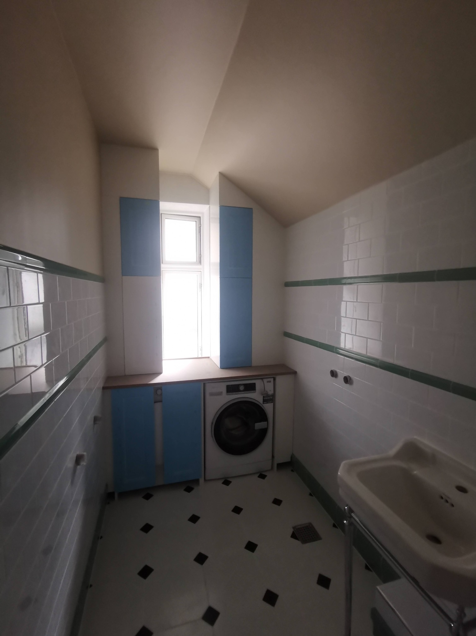 Ukończona łazienka na poddaszu z biało-niebieskimi szafkami, pralką zabudowaną pod blatem, białą glazurą z zielonym pasem i podłogą w czarno-białą szachownicę.