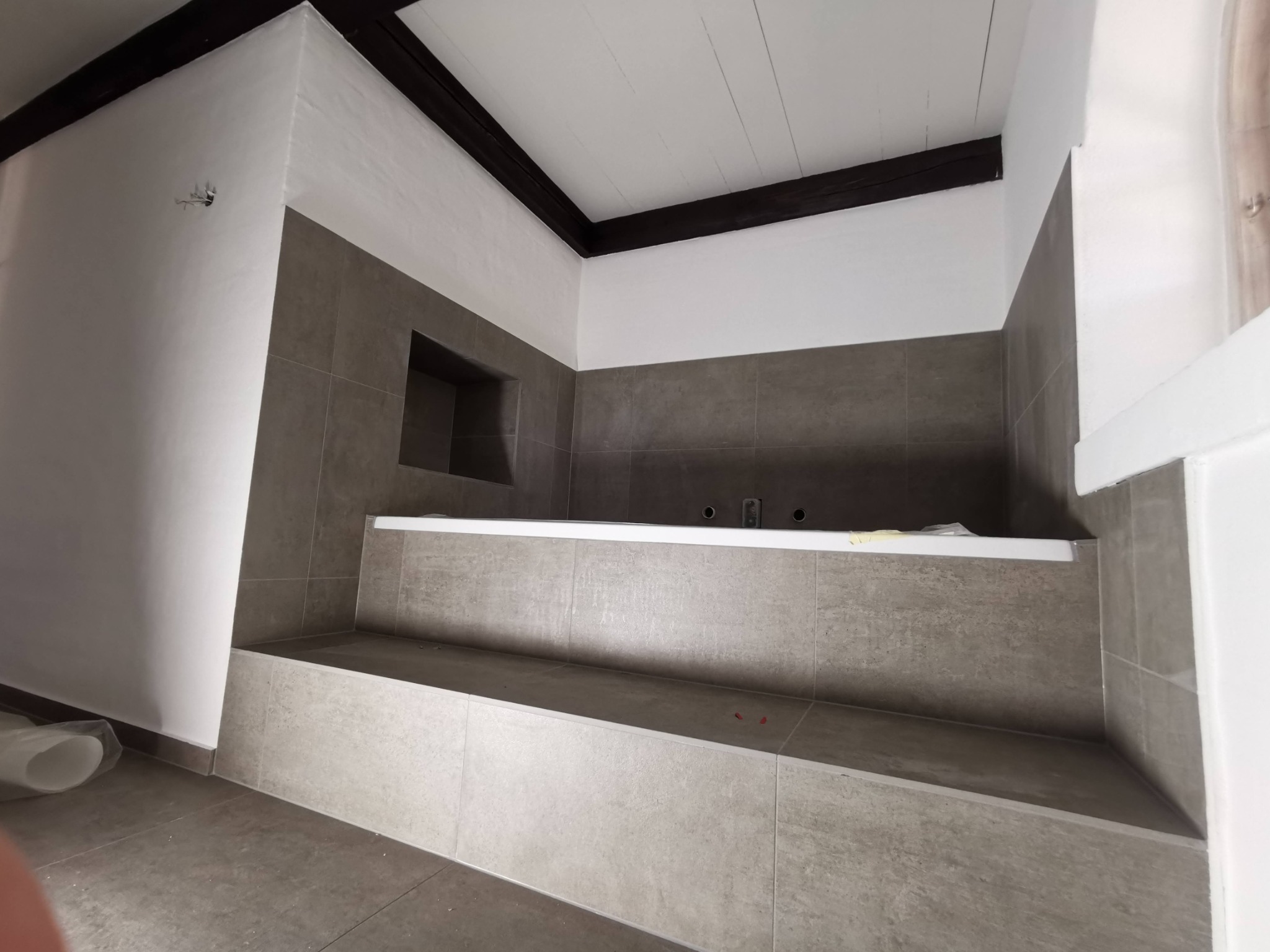 Wnętrze łazienki w trakcie prac wykończeniowych, widoczna wanna obudowana płytkami w odcieniach szarości, z ciemnymi belkami pod sufitem, wnęka w ścianie nad wanną.