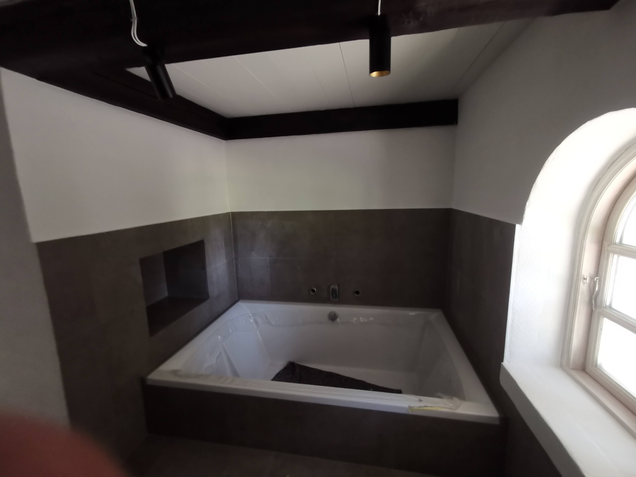 Wanna w trakcie montażu, zabezpieczona folią, w łazience z ciemnymi płytkami i drewnianymi belkami sufitowymi, widoczne przygotowane otwory na armaturę.