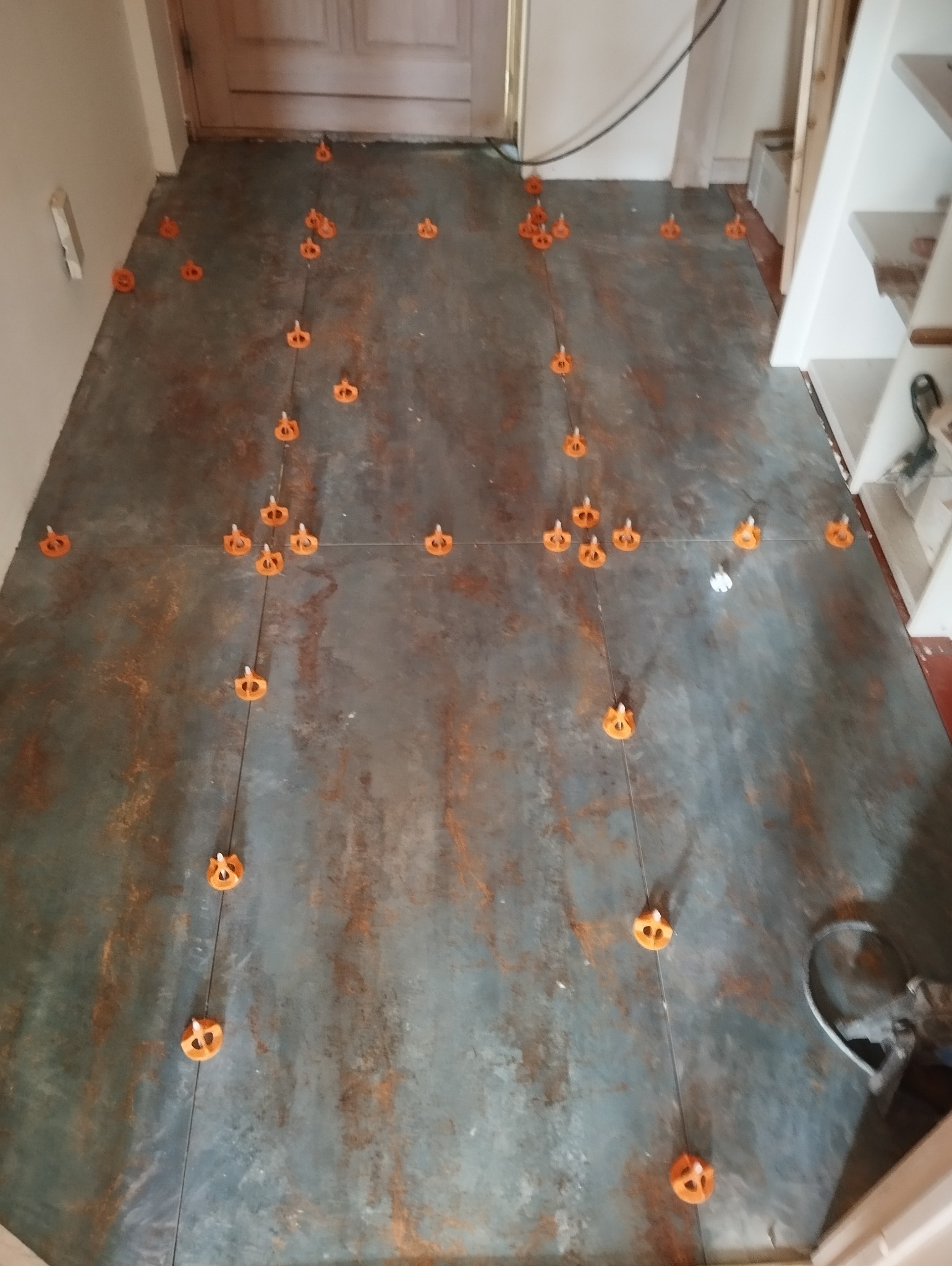 Podłoga z szarych płytek imitujących rdzę, z pomarańczowymi klipsami poziomującymi i cienkimi sznurkami do fugowania, widoczne fragmenty listew przypodłogowych i drzwi.