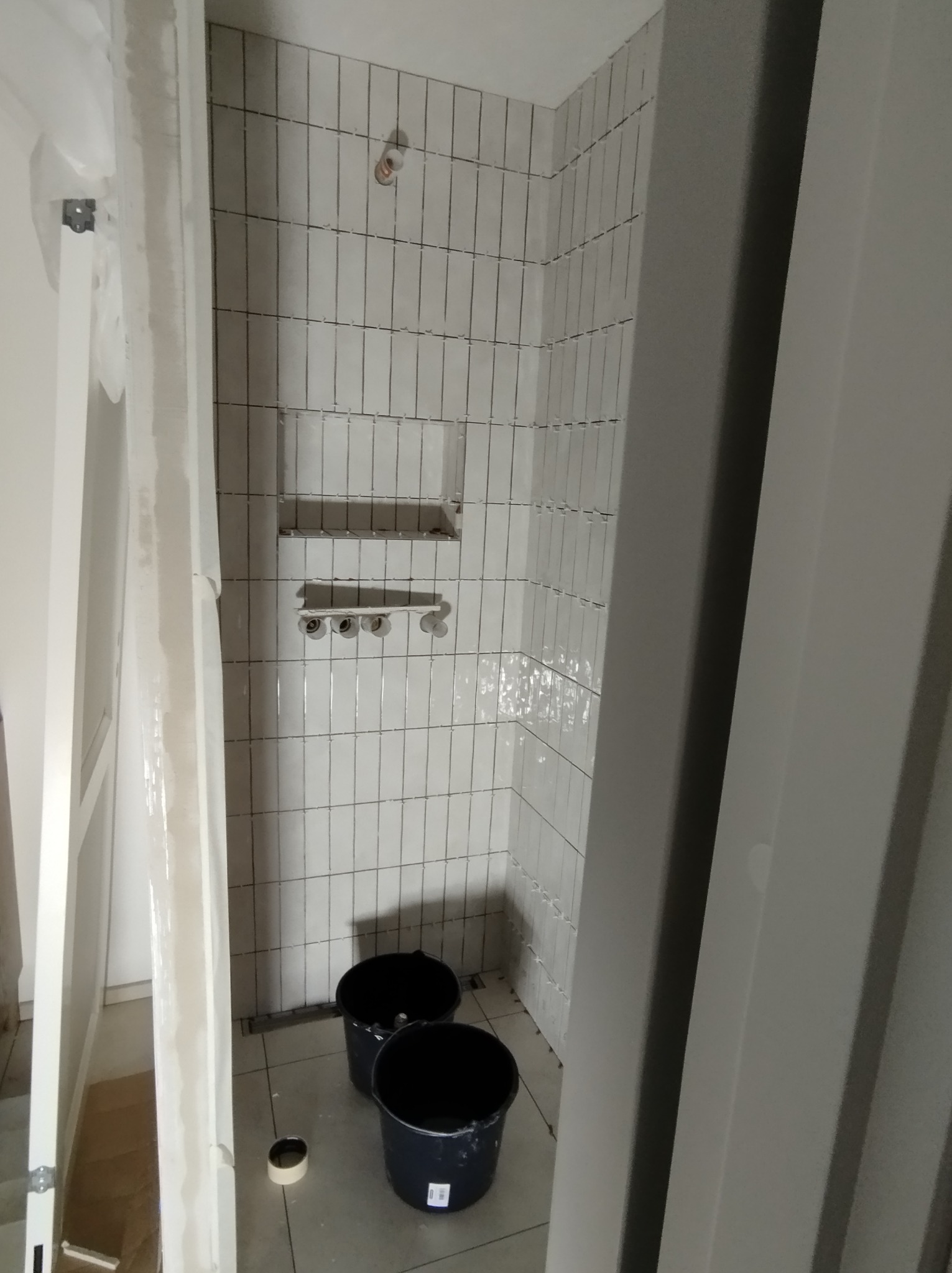 Wnętrze łazienki w trakcie prac, widoczne pionowo ułożone białe płytki, przygotowane półki, dwa czarne wiadra na podłodze i elementy ościeżnicy drzwi.