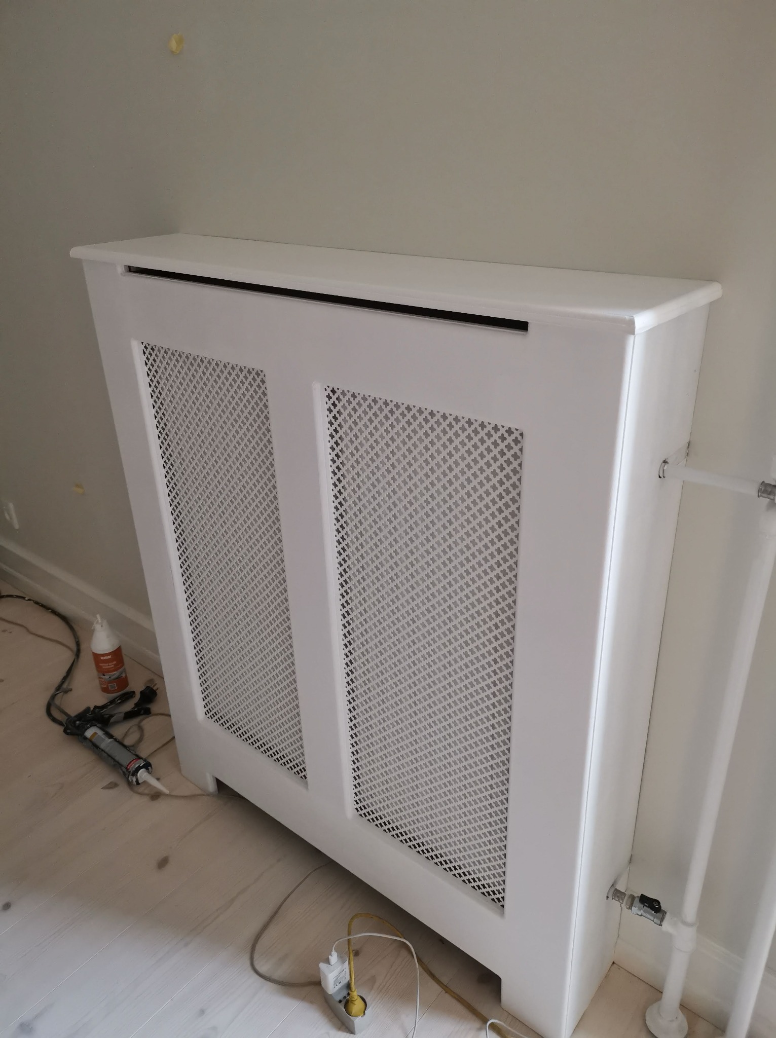 Biała, ażurowa obudowa grzejnika zamontowana przy ścianie, widoczne narzędzia montażowe i kabel zasilający na podłodze.