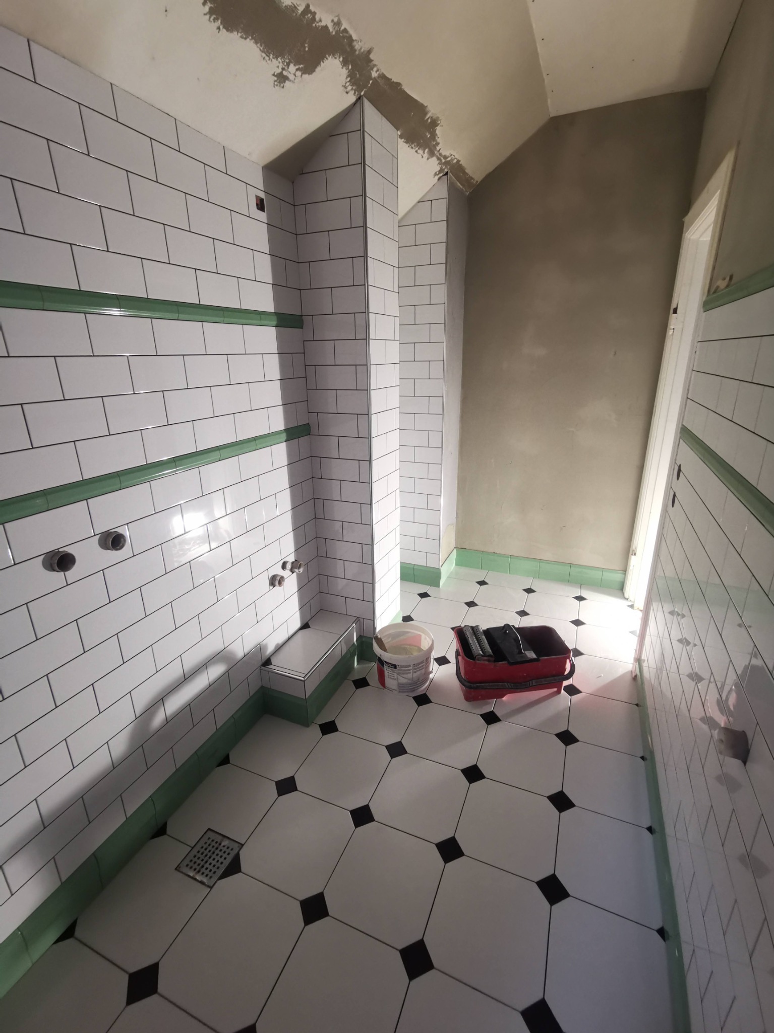 Wnętrze łazienki w trakcie prac wykończeniowych: białe płytki typu metro z zielonym akcentem, czarno-biała podłoga w karo, widoczne narzędzia i wiadro z zaprawą, surowy sufit z pozostałościami...