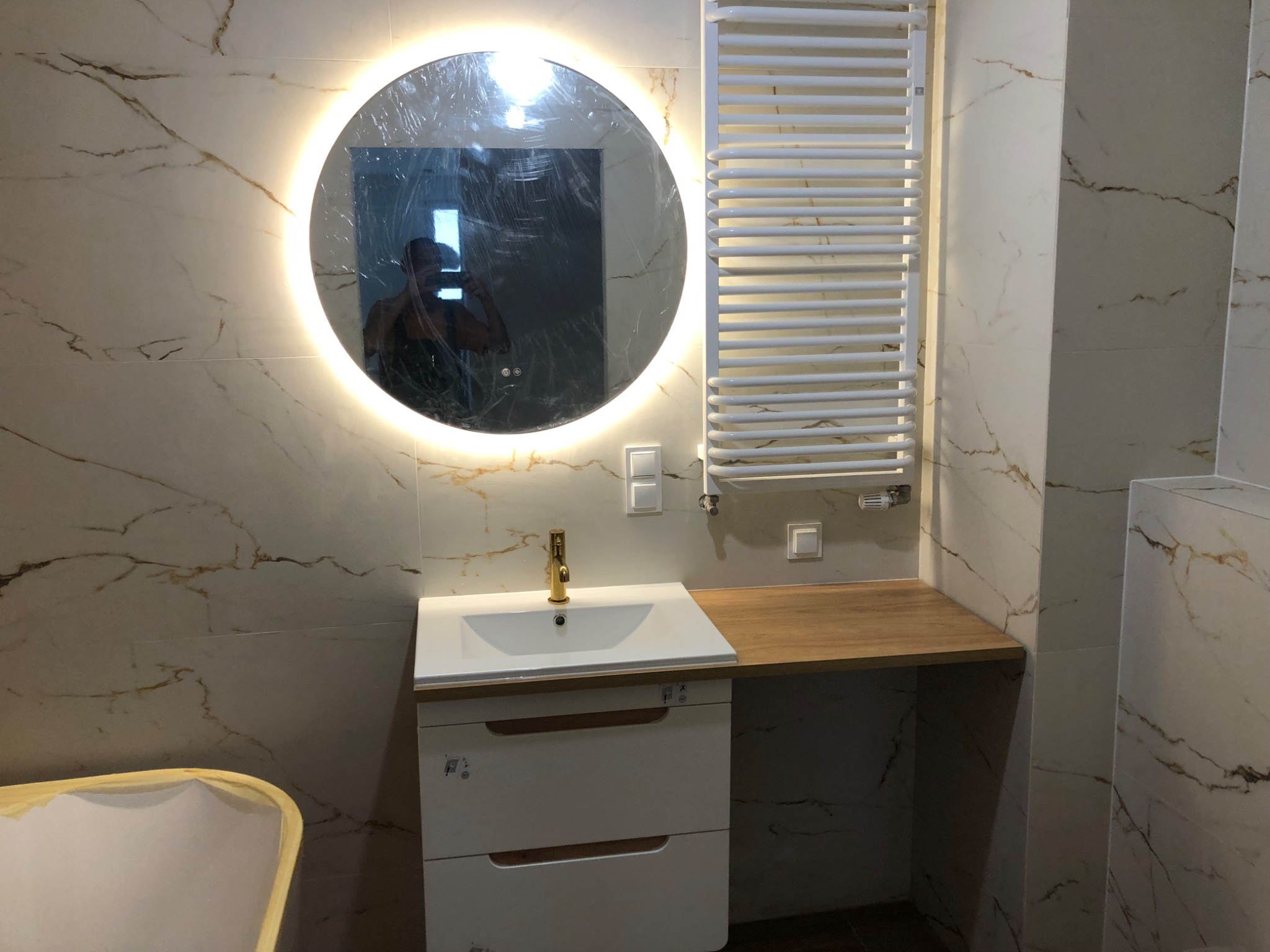 Nowoczesna łazienka z umywalką nablatową, okrągłym lustrem z podświetleniem LED i grzejnikiem drabinkowym na marmurowej ścianie, widoczne folie ochronne na lustrze.