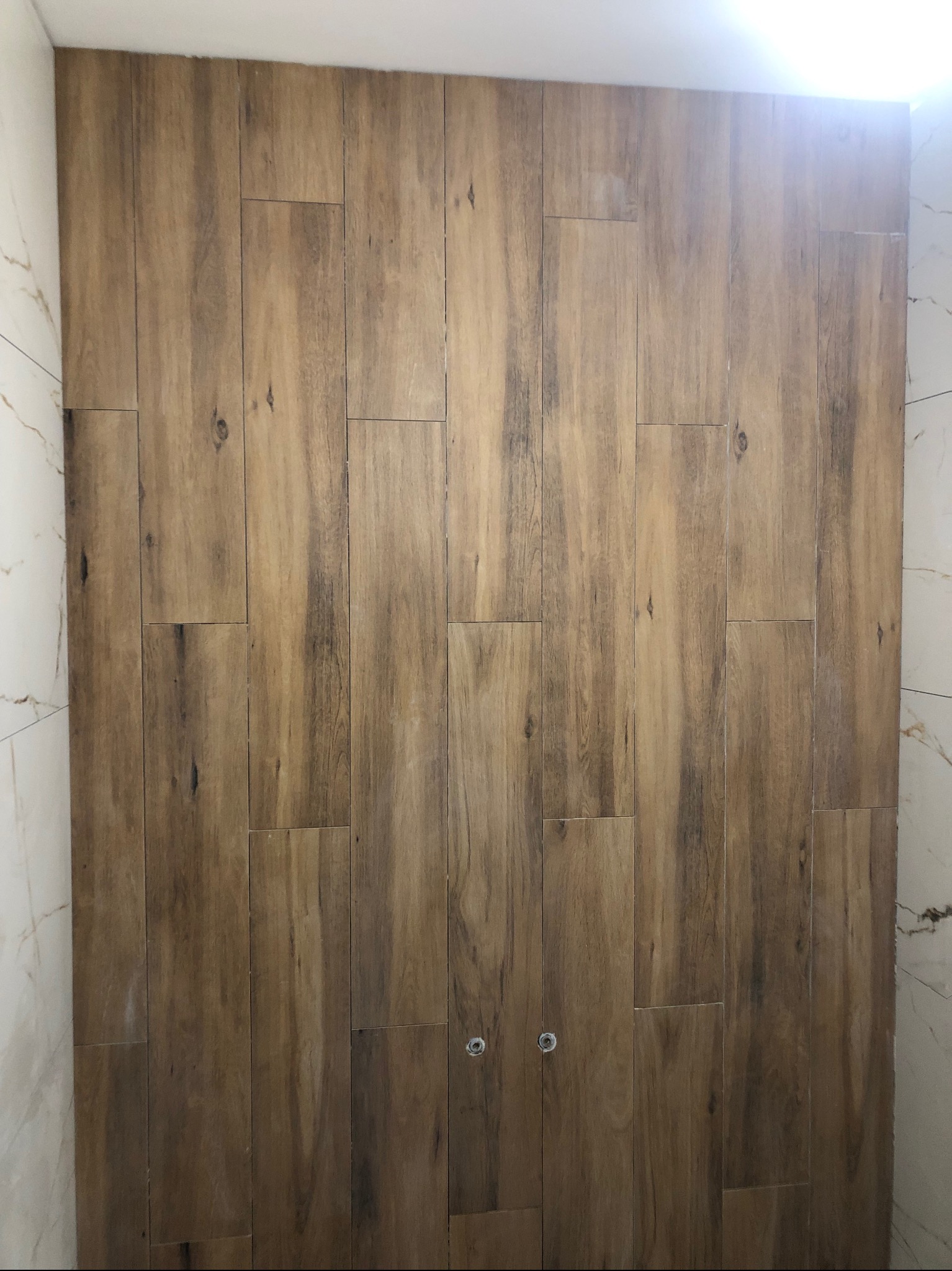 Ściana w łazience wyłożona podłużnymi płytkami imitującymi drewno, z widocznymi dwoma chromowanymi elementami instalacji, sąsiadująca z białą ścianą wyłożoną marmurkowymi płytkami.