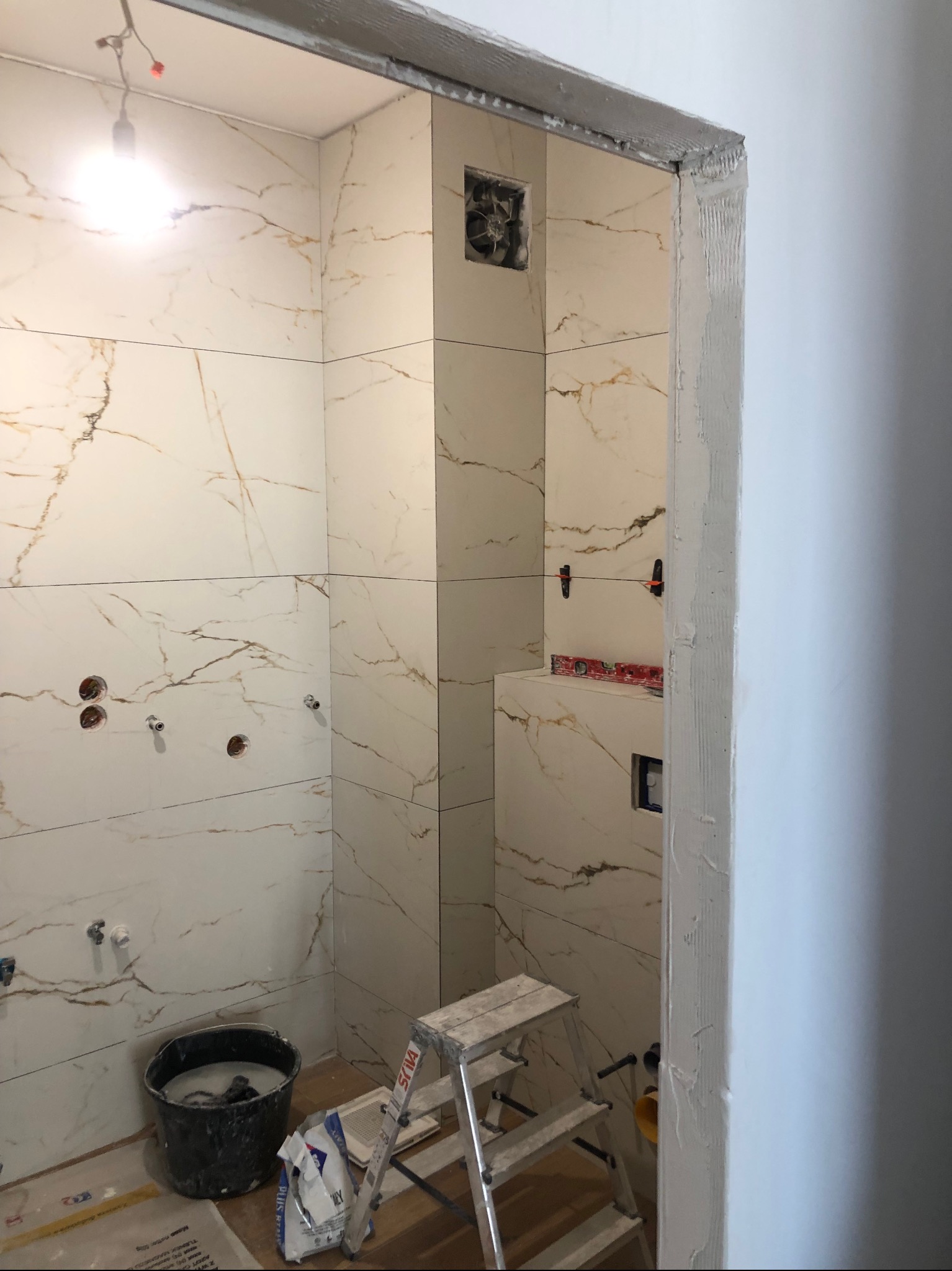 Wnętrze łazienki w trakcie układania białych płytek z marmurowym wzorem, widoczna drabina, wiadro z zaprawą, poziomica i elementy instalacji.
