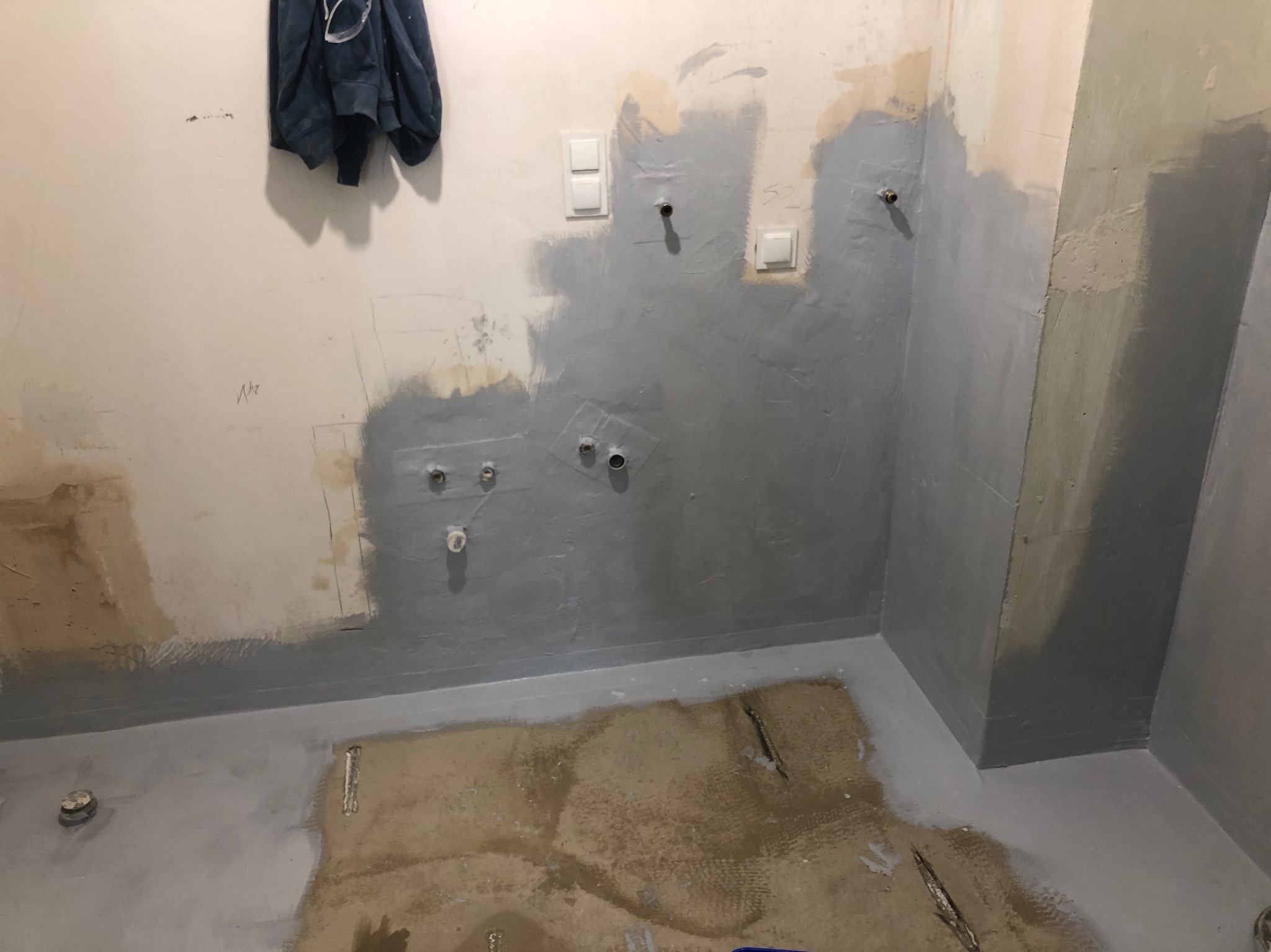 Przygotowanie ścian i podłogi do remontu łazienki: częściowo pomalowane ściany na szaro, widoczne wystające rury, przełączniki elektryczne i zabezpieczona podłoga przed położeniem płytek.