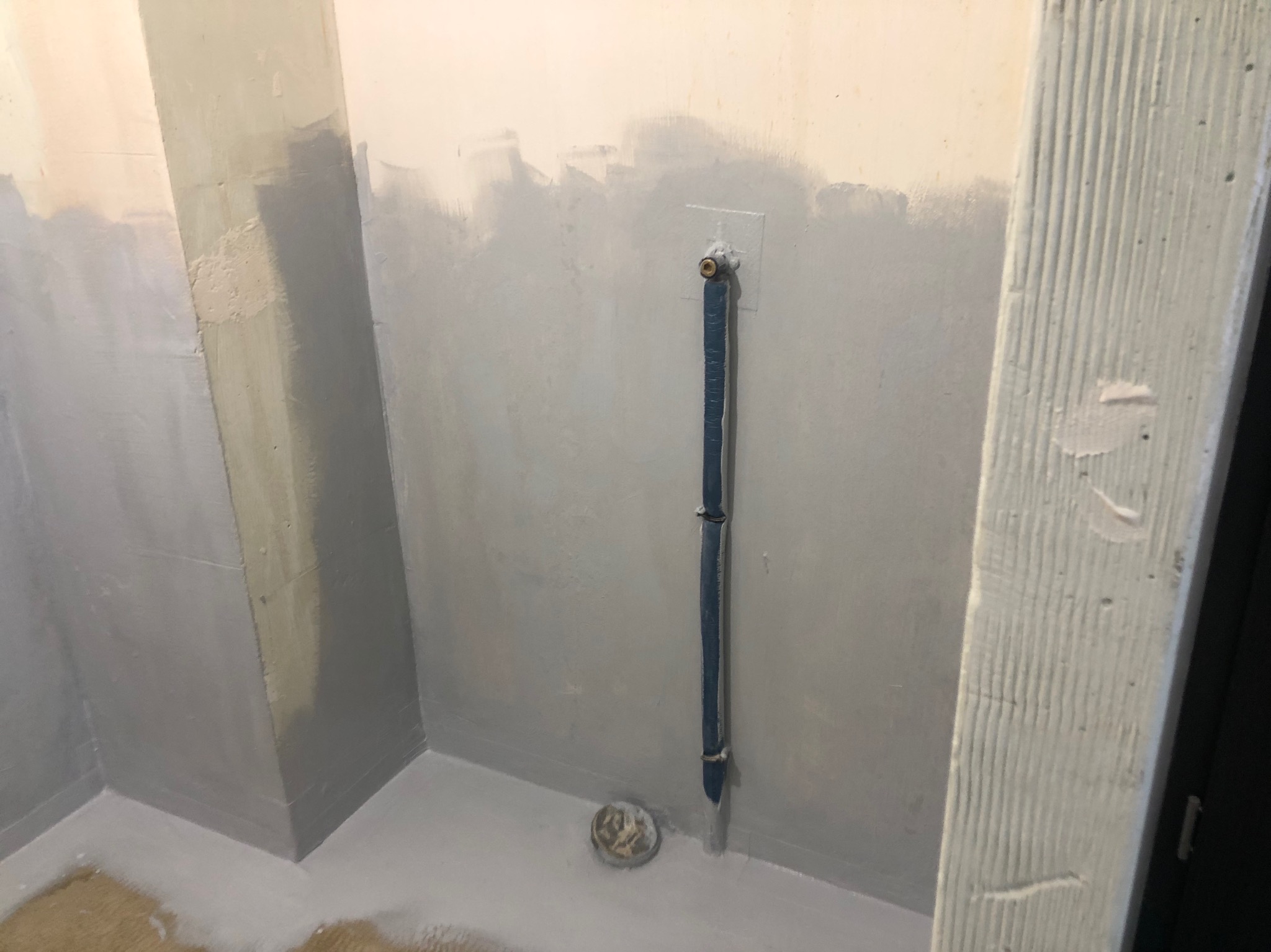 Przygotowana do dalszych prac remontowych ściana w łazience z widocznym pionem instalacji wodnej i odpływem w podłodze, zabezpieczona folią w płynie.