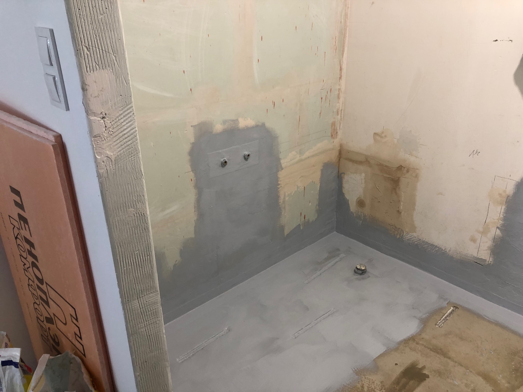 Przygotowanie do hydroizolacji w narożniku łazienki: szare uszczelnienie na ścianach i podłodze, widoczne wystające rury, obok płyta styropianowa.