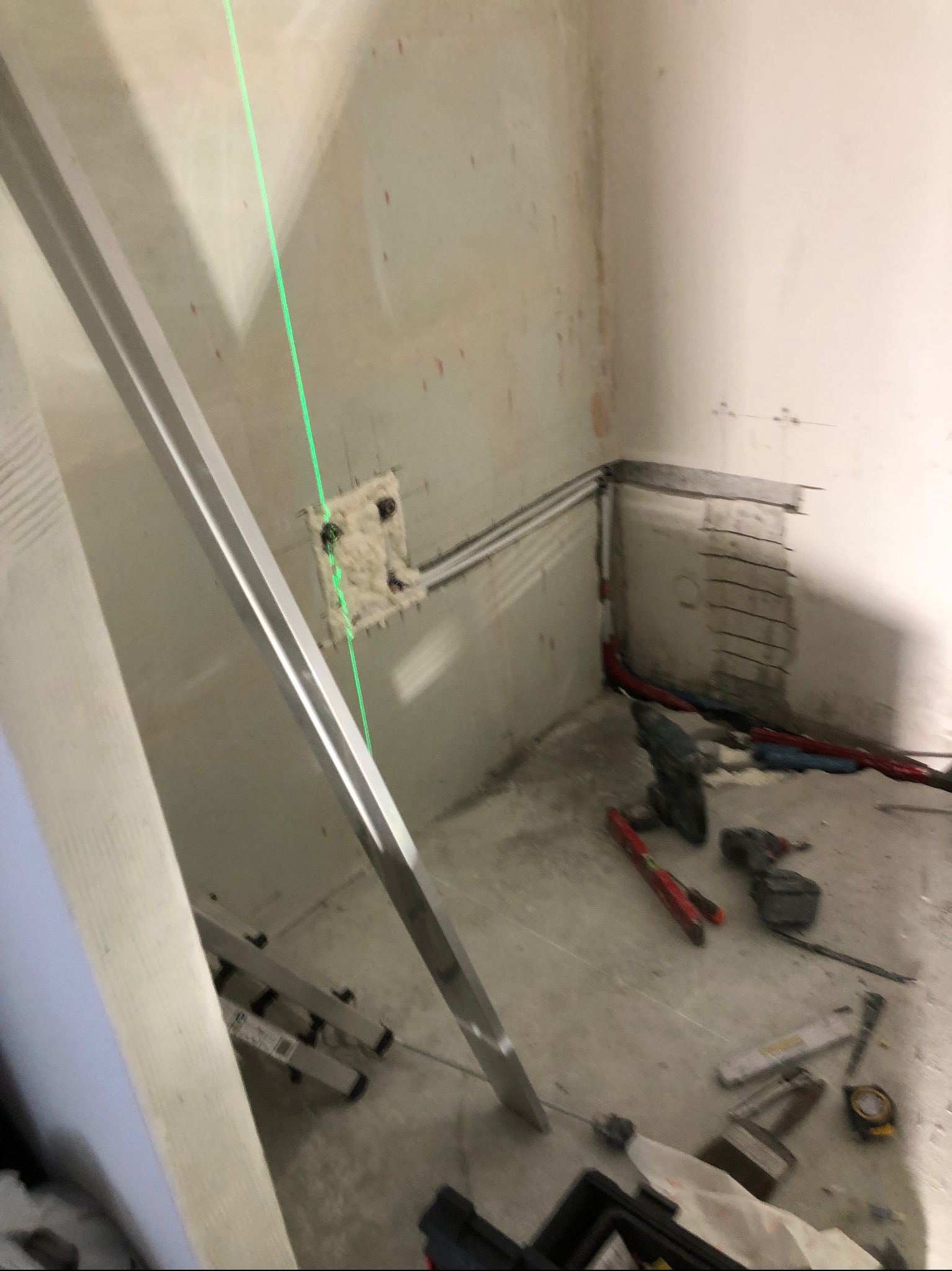 Wnętrze łazienki w trakcie remontu, widoczny laser krzyżowy wyznaczający poziom, przewody instalacji elektrycznej zabezpieczone pianką montażową, narzędzia na podłodze, ściany przygotowane...