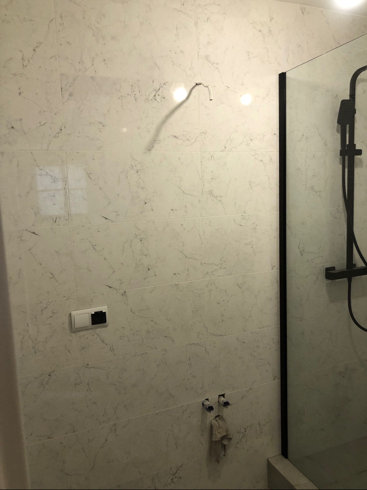 Ściana łazienki wyłożona białymi płytkami imitującymi marmur, z widoczną czarną kabiną prysznicową, wystającymi rurami i wiszącym ręcznikiem, w trakcie prac wykończeniowych.