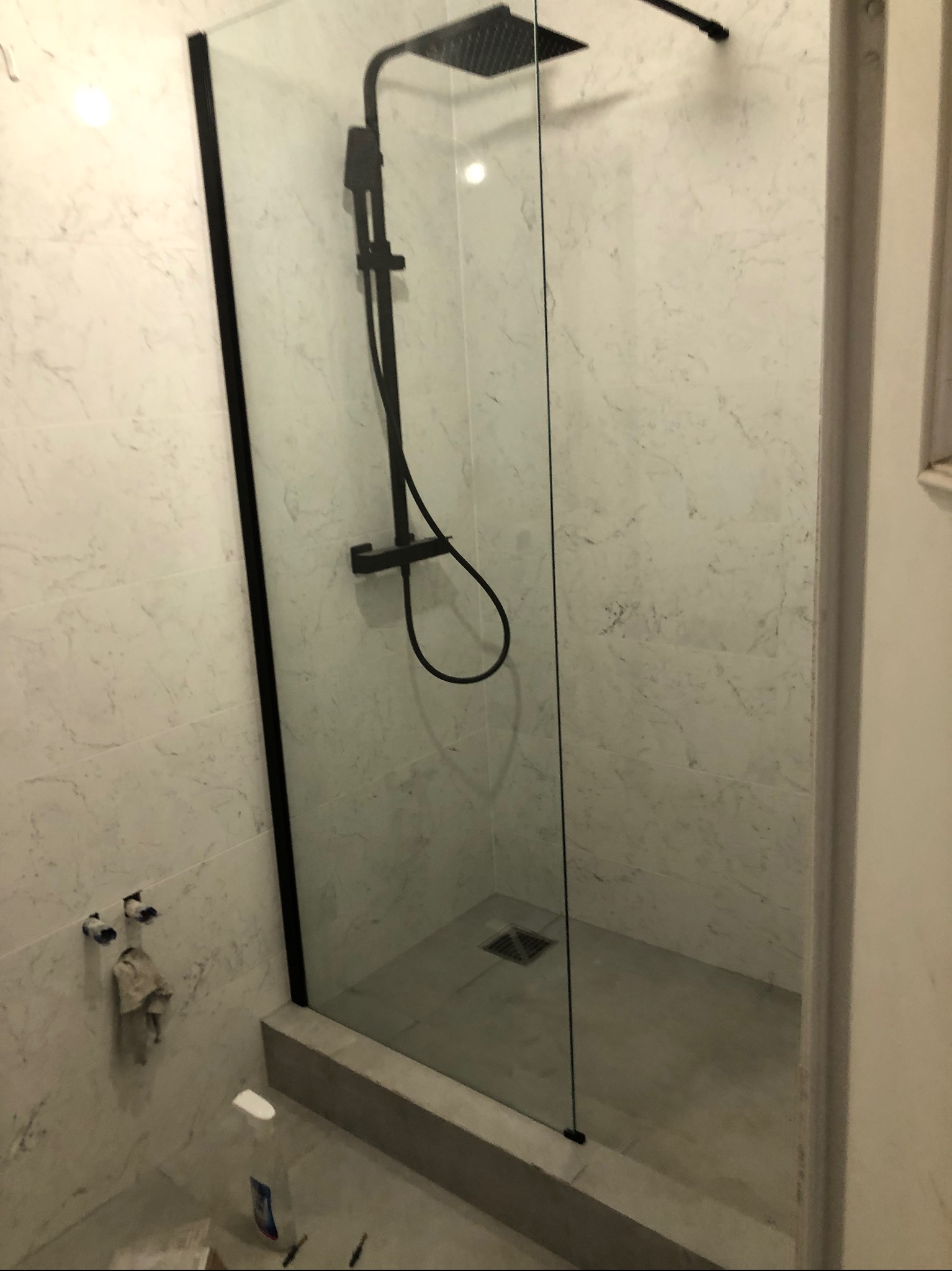 Nowoczesna kabina prysznicowa typu walk-in z czarną armaturą na tle marmurowych płytek, widoczne niedokończone przyłącza i butelka środka czyszczącego.