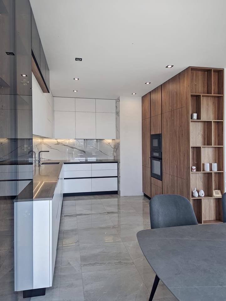 Nowoczesna kuchnia z białymi szafkami z połyskiem, blatem imitującym kamień, drewnianą zabudową z piekarnikiem i otwartymi półkami oraz szarą podłogą z płytek ceramicznych.