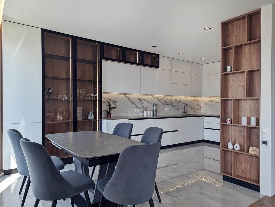 Nowoczesna kuchnia z białymi szafkami, marmurowym backsplashiem, drewnianymi akcentami i szarymi krzesłami przy stole, oświetlona listwami LED pod szafkami.