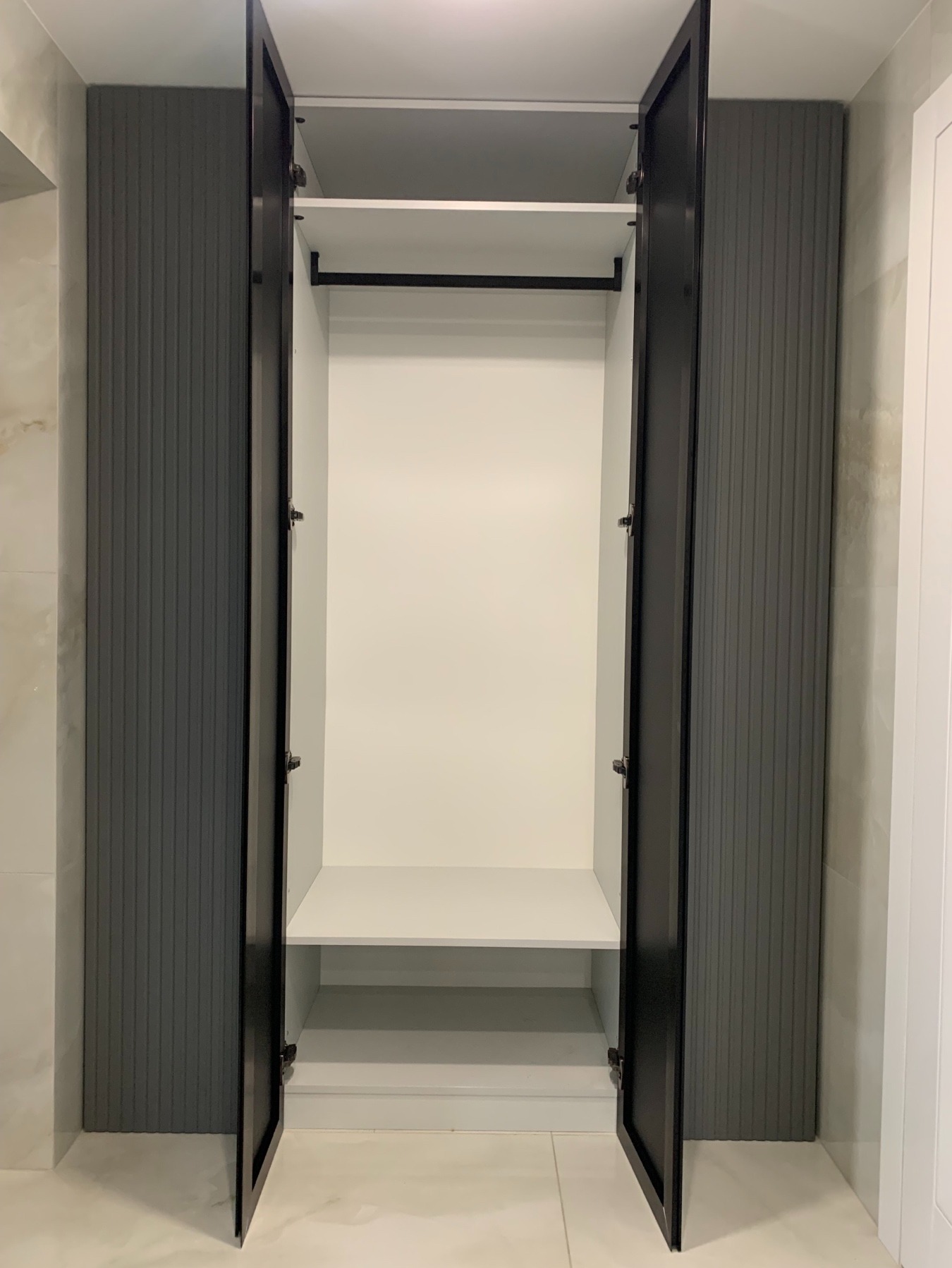Wnętrze szafy wnękowej z otwartymi drzwiami, widoczne półki i drążek na wieszaki, szare panele boczne z pionowymi żłobieniami, czarne obramowanie drzwi, białe ściany i podłoga.