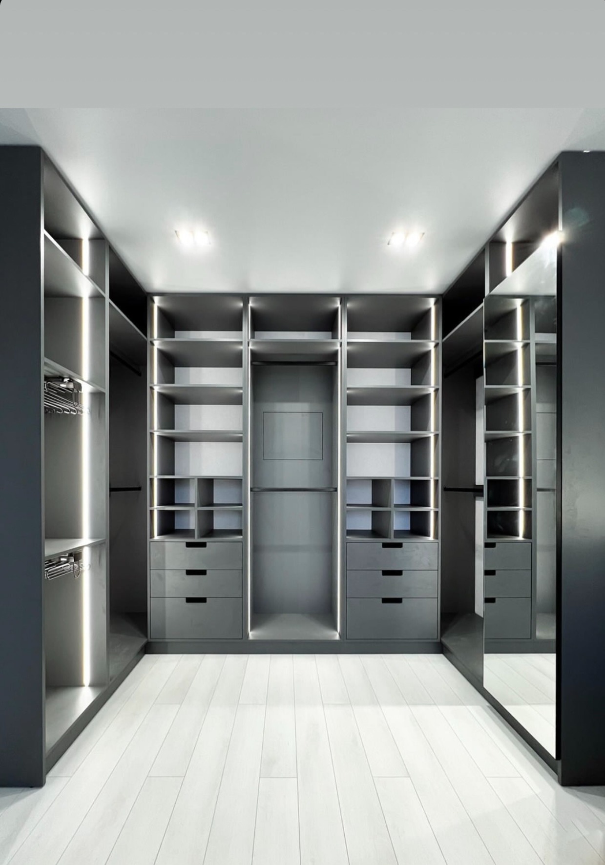 Nowoczesna, szara garderoba z oświetleniem LED wbudowanym w półki i szuflady, z jasną drewnianą podłogą i lustrem na ścianie.