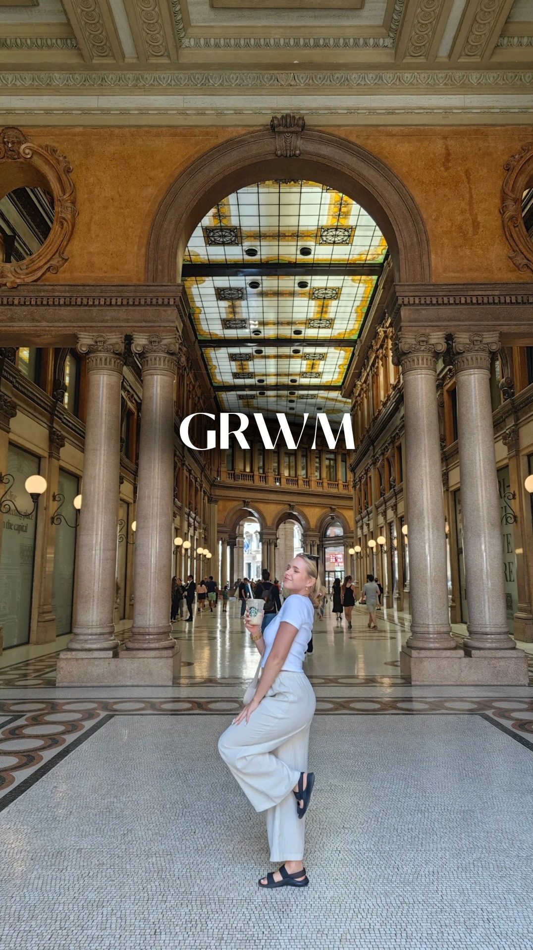 Kobieta pozuje w galerii handlowej z kolumnami, trzymając napój. Nałożony napis GRWM.