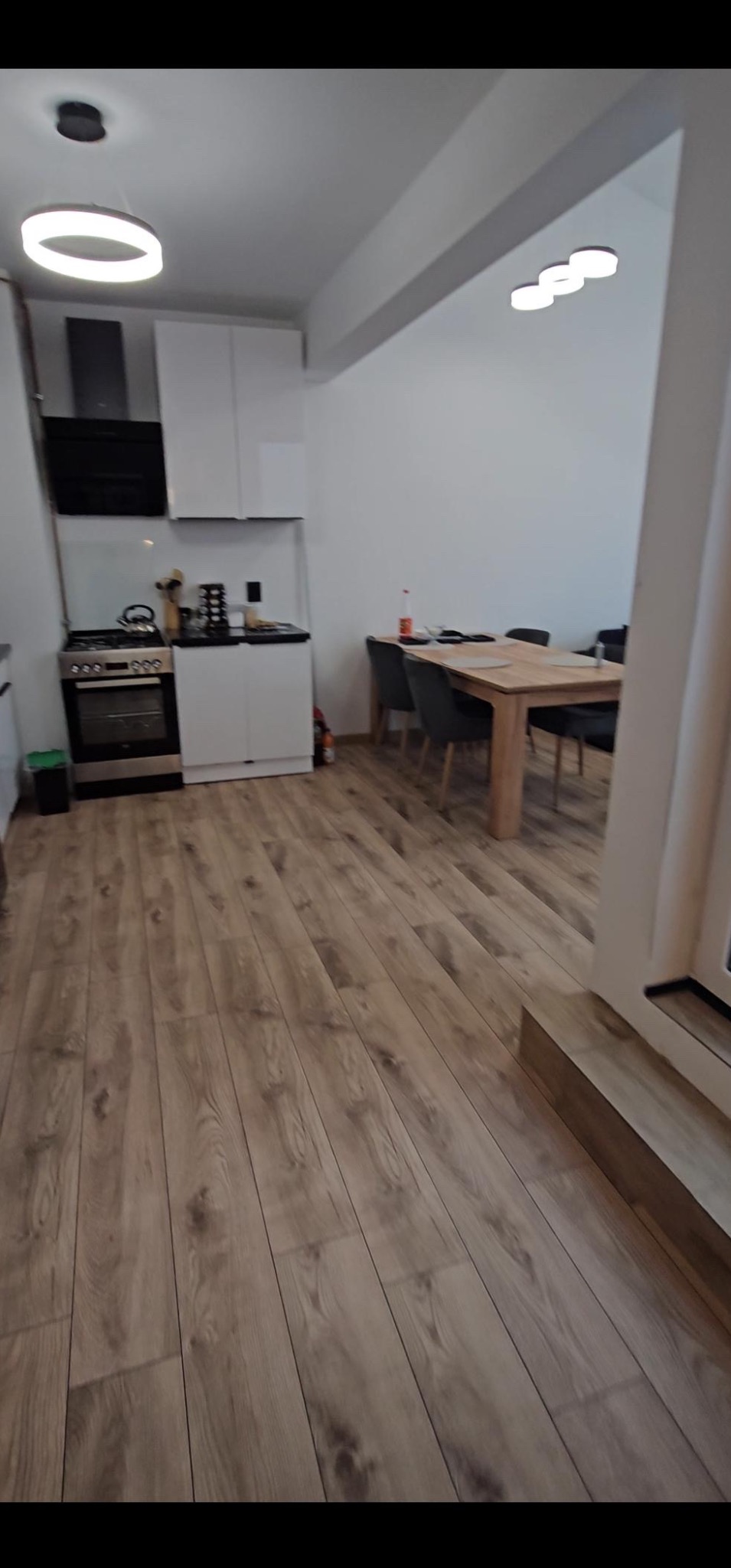 Wykończone wnętrze mieszkania z jasnymi panelami podłogowymi o wzorze drewna, widok na kuchnię z białymi szafkami i stół jadalny z krzesłami w tle.