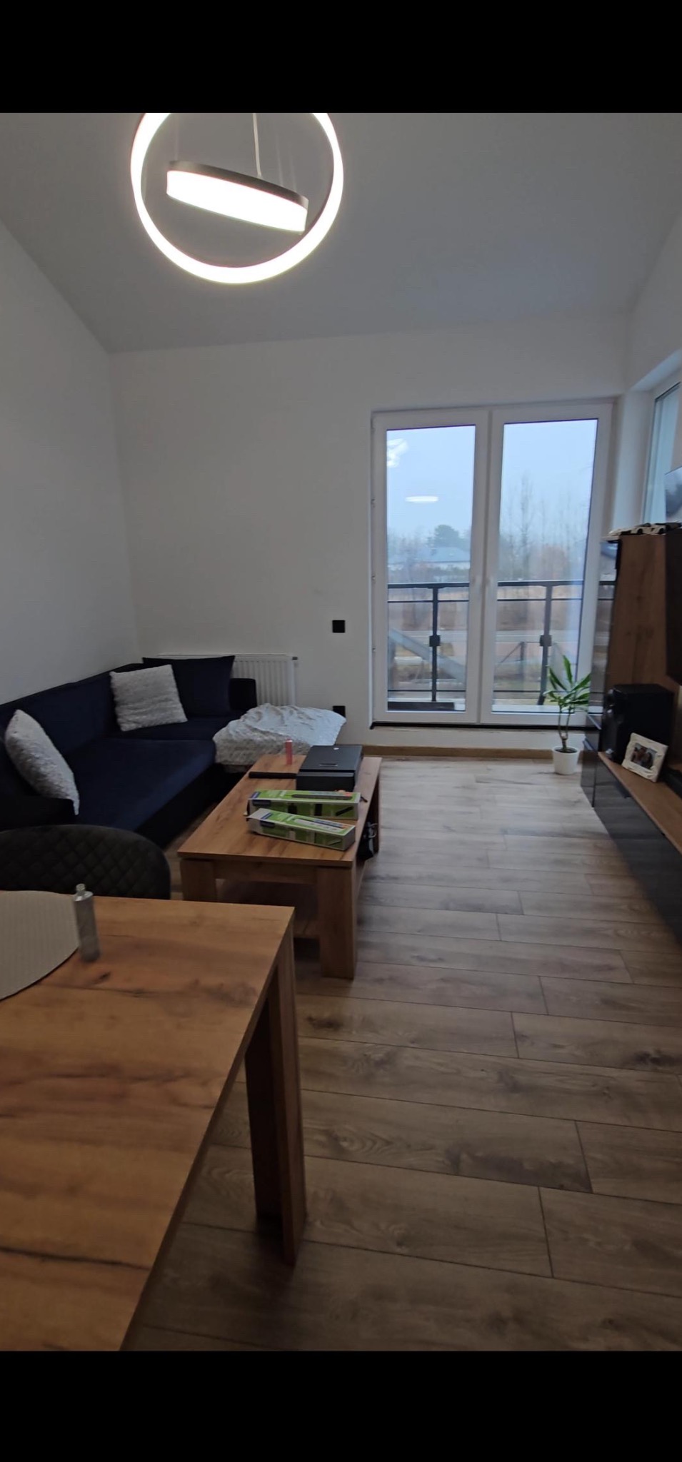 Salon w mieszkaniu z drewnianą podłogą, narożnikiem i widokiem na balkon. Na stole widoczne opakowania po listwach przypodłogowych.