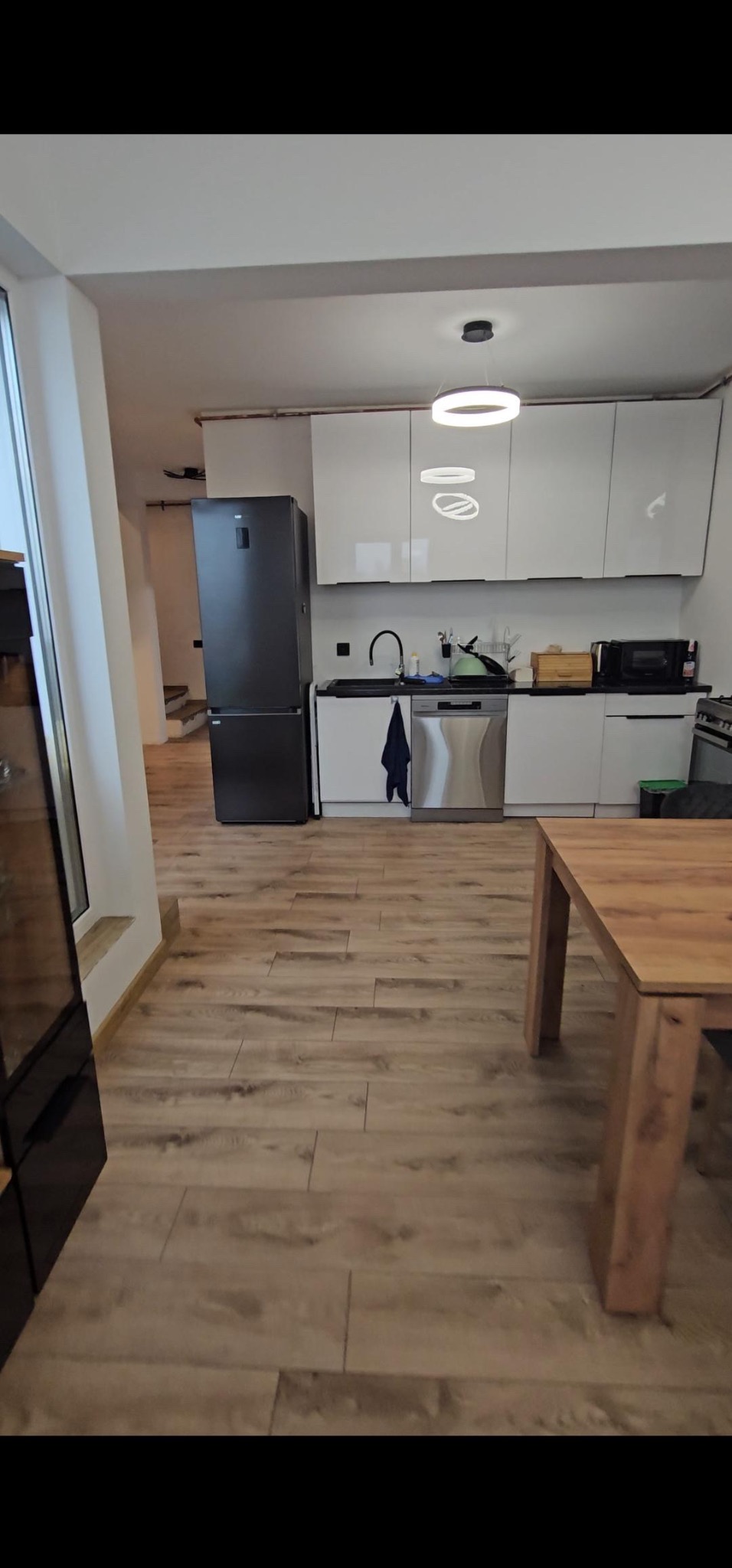 Kuchenne wnętrze z drewnianą podłogą z paneli, widok na białe szafki, czarną lodówkę i fragment drewnianego stołu.