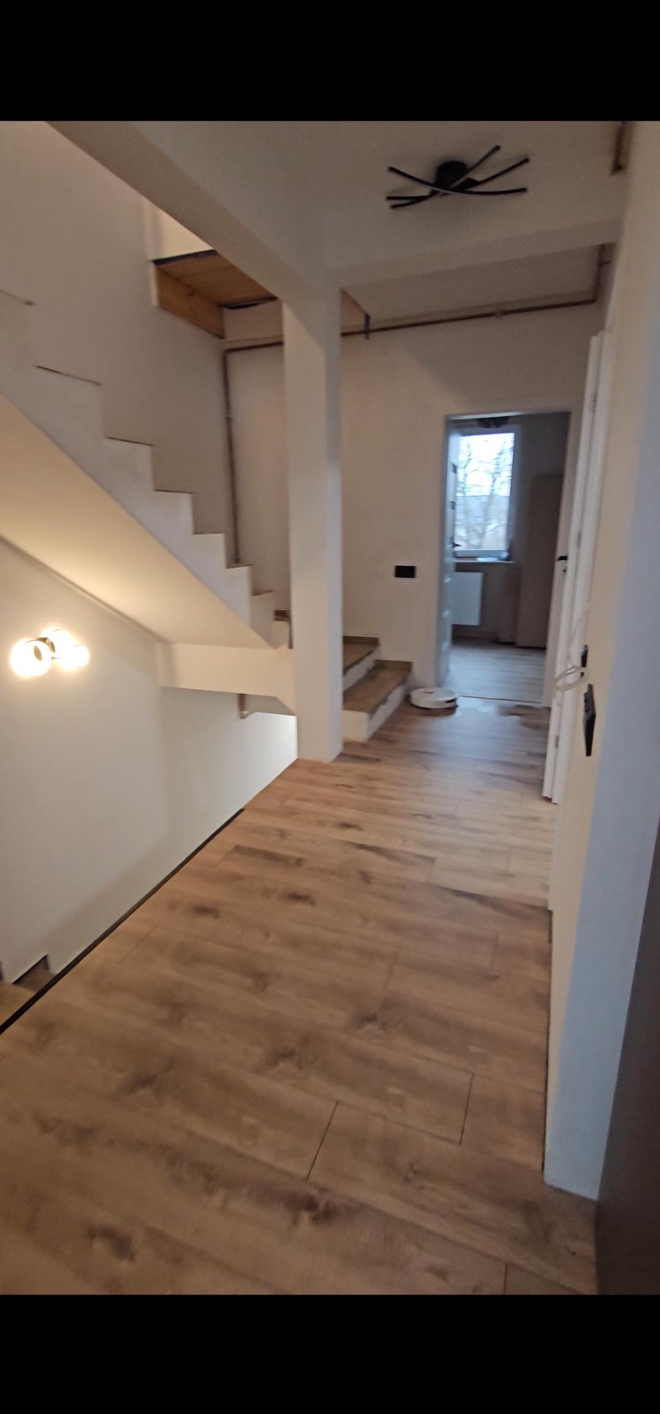 Nowoczesne wnętrze domu z drewnianą podłogą, białymi ścianami i schodami. Widoczny odkurzacz automatyczny. Zwraca uwagę minimalistyczna lampa sufitowa.